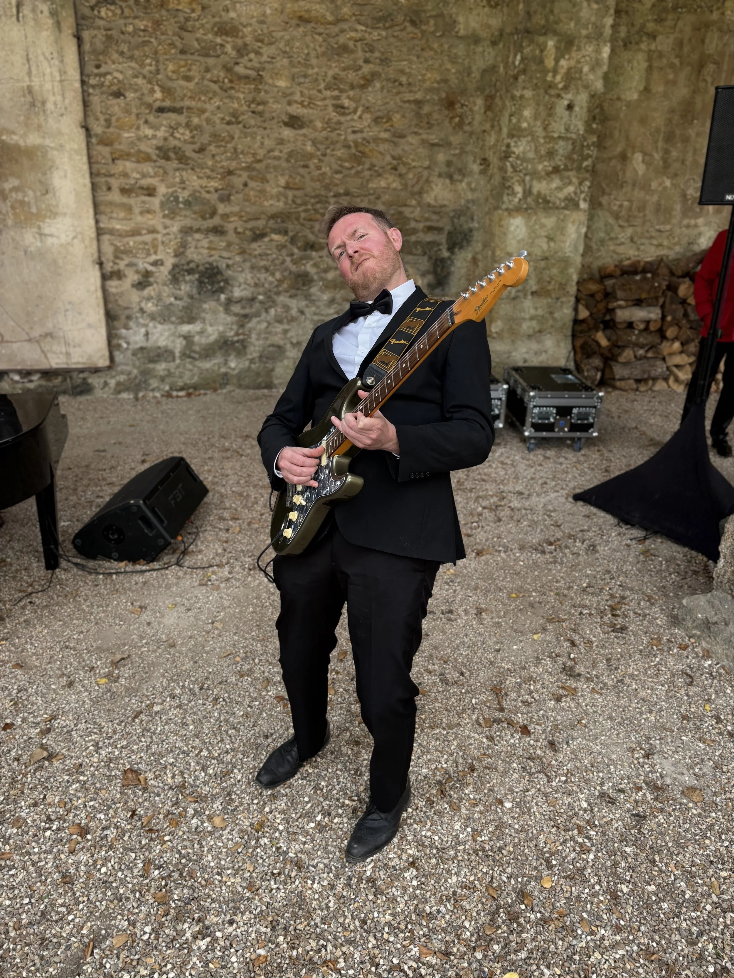 A man dressed in a black tuxedo with a bowtie is holding a guitar and making a face with his head tilted to one side. The background features a textured stone wall, a pile of firewood, and some music equipment.