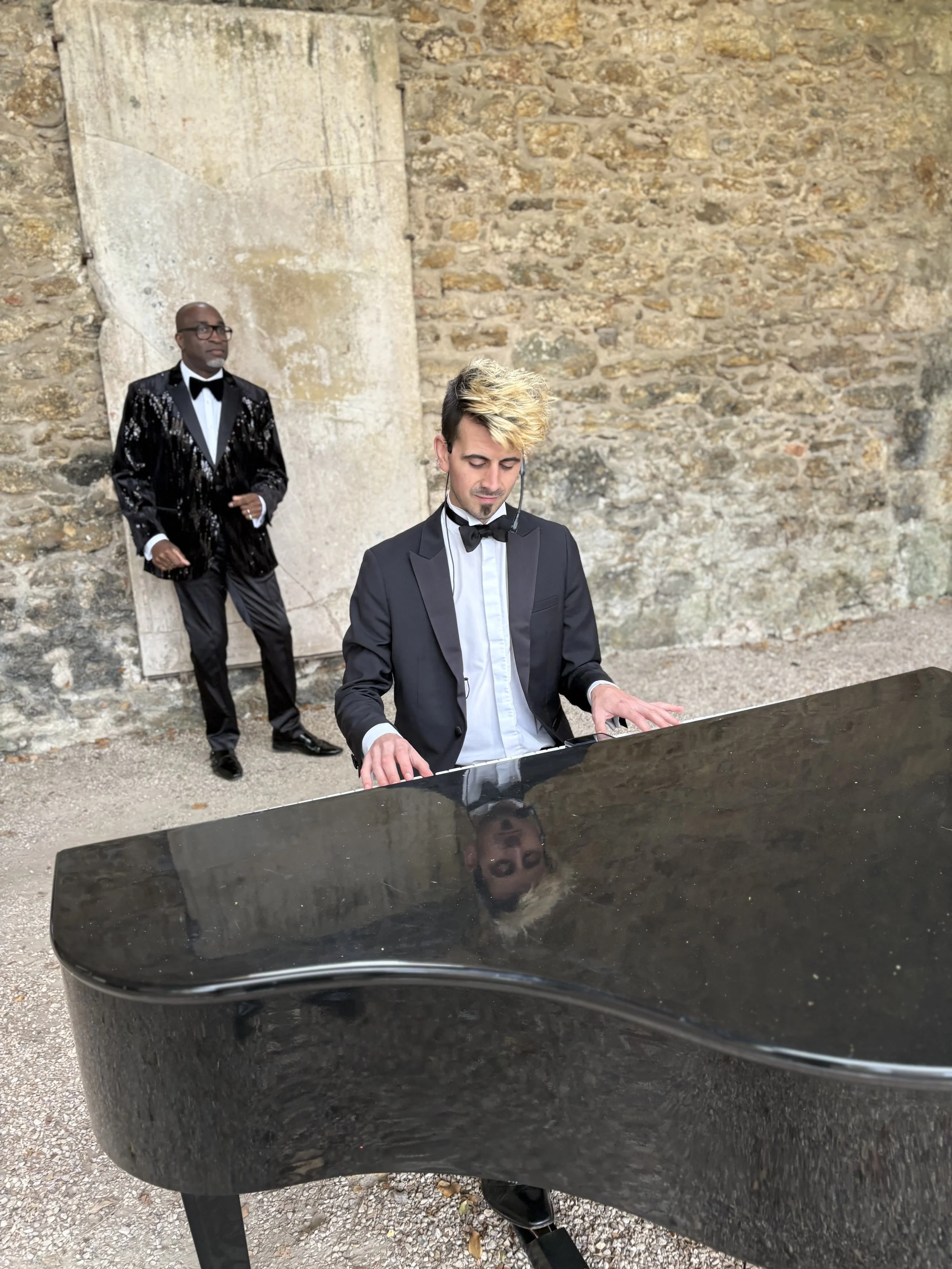 A man in a formal tuxedo playing a black grand piano outdoors, with a second man in a tuxedo standing in the background against a stone wall.
