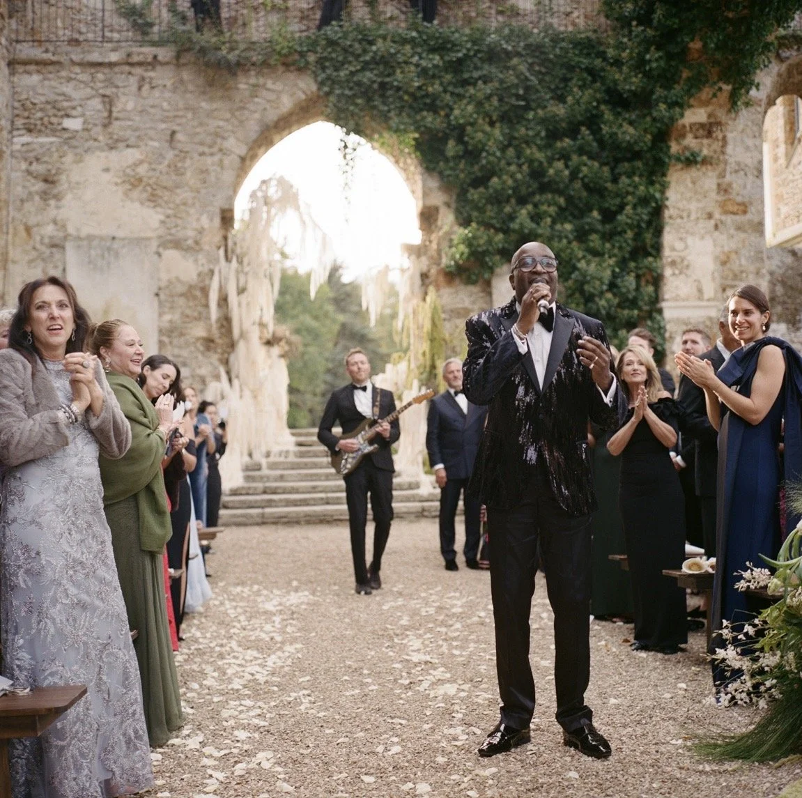 A man in a black tuxedo with a shiny black jacket is singing into a microphone at a wedding reception, surrounded by guests clapping and smiling under an archway with old stone walls and greenery.