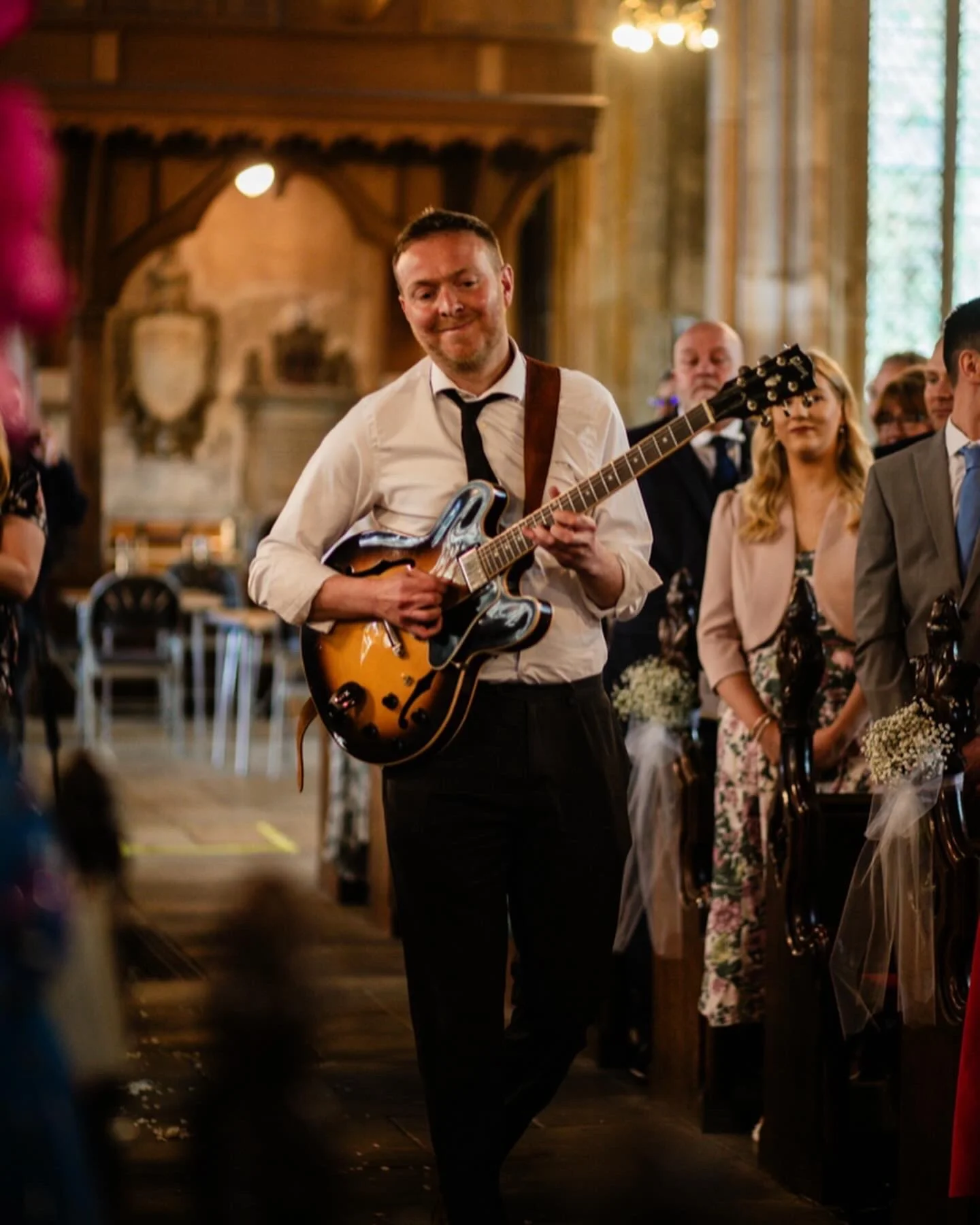 Groom playing guitar at wedding reception in a decorated venue with guests seated at tables.