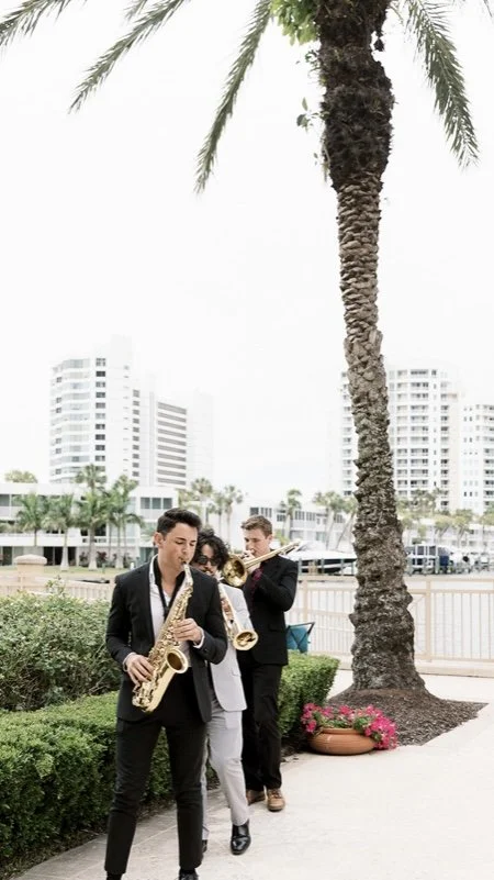 Three musicians playing saxophones and a trumpet outdoors near a palm tree with city buildings in the background.