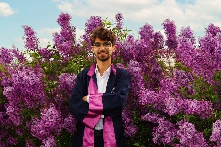 Un jeune homme souriant en tenue de graduation, debout devant un enclos de buissons de sassafras en fleurs violettes, sous un ciel partiellement nuageux.
