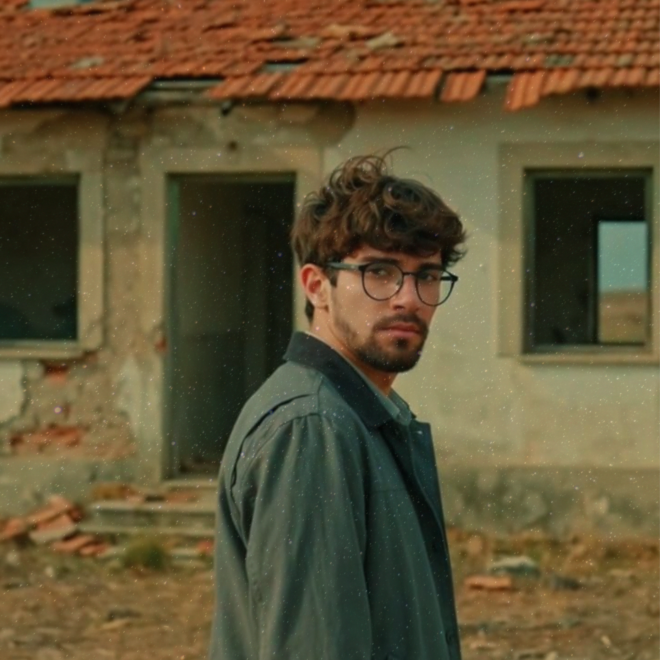 Un jeune homme avec des lunettes, des cheveux bruns et bouclés, portant une veste sombre, se tient devant une maison avec un toit en tuiles rouges et des fenêtres, dans un environnement extérieur.