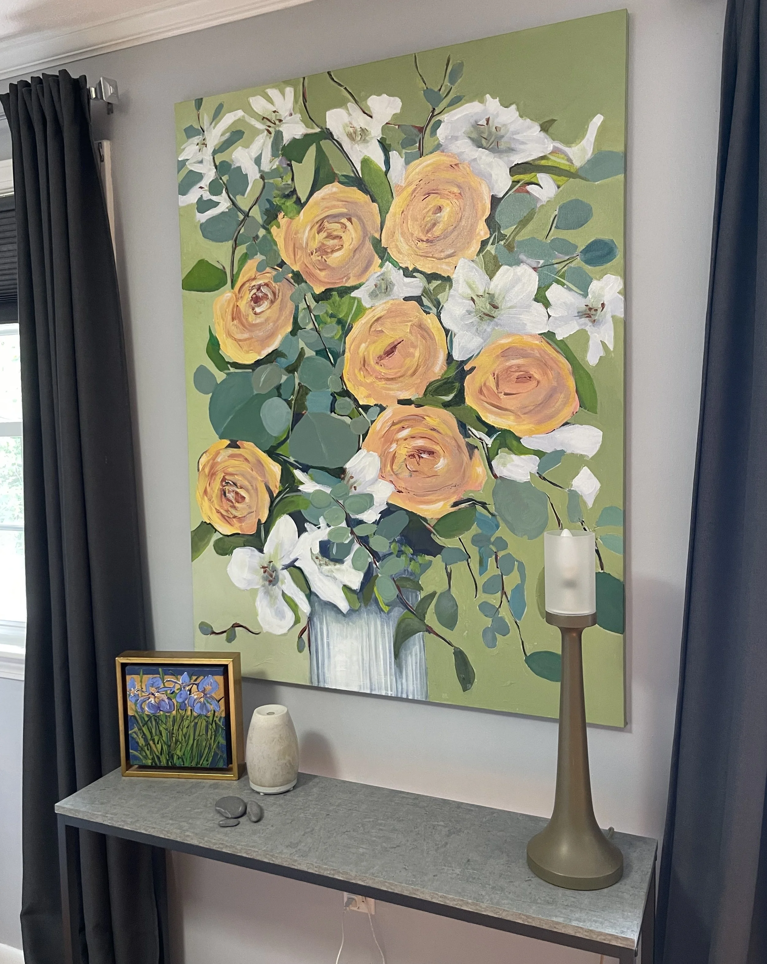 Interior of a room with a large floral painting of Yellow Roses on the wall, a smaller floral artwork on the table, a beige table lamp, a white vase, and some stones on the table.