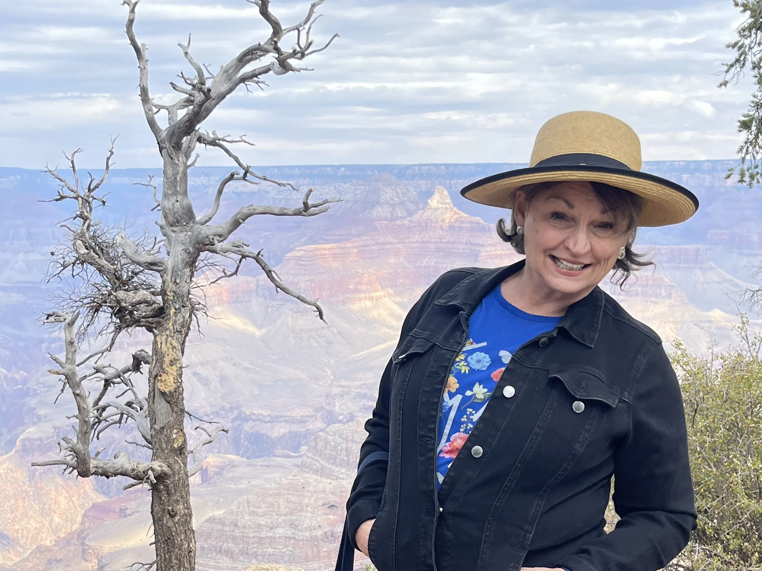 A smiling woman wearing a wide-brimmed straw hat, a blue floral T-shirt, and a black jacket standing near a leafless tree with the Grand Canyon in the background. The sky is mostly cloudy.