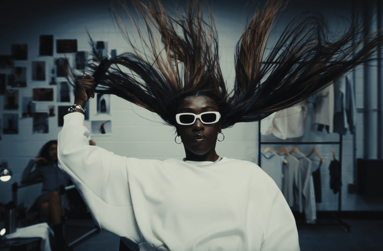 A woman with long flowing hair wearing white sunglasses and a white oversized top, standing in a photo studio with photos on the wall behind her.