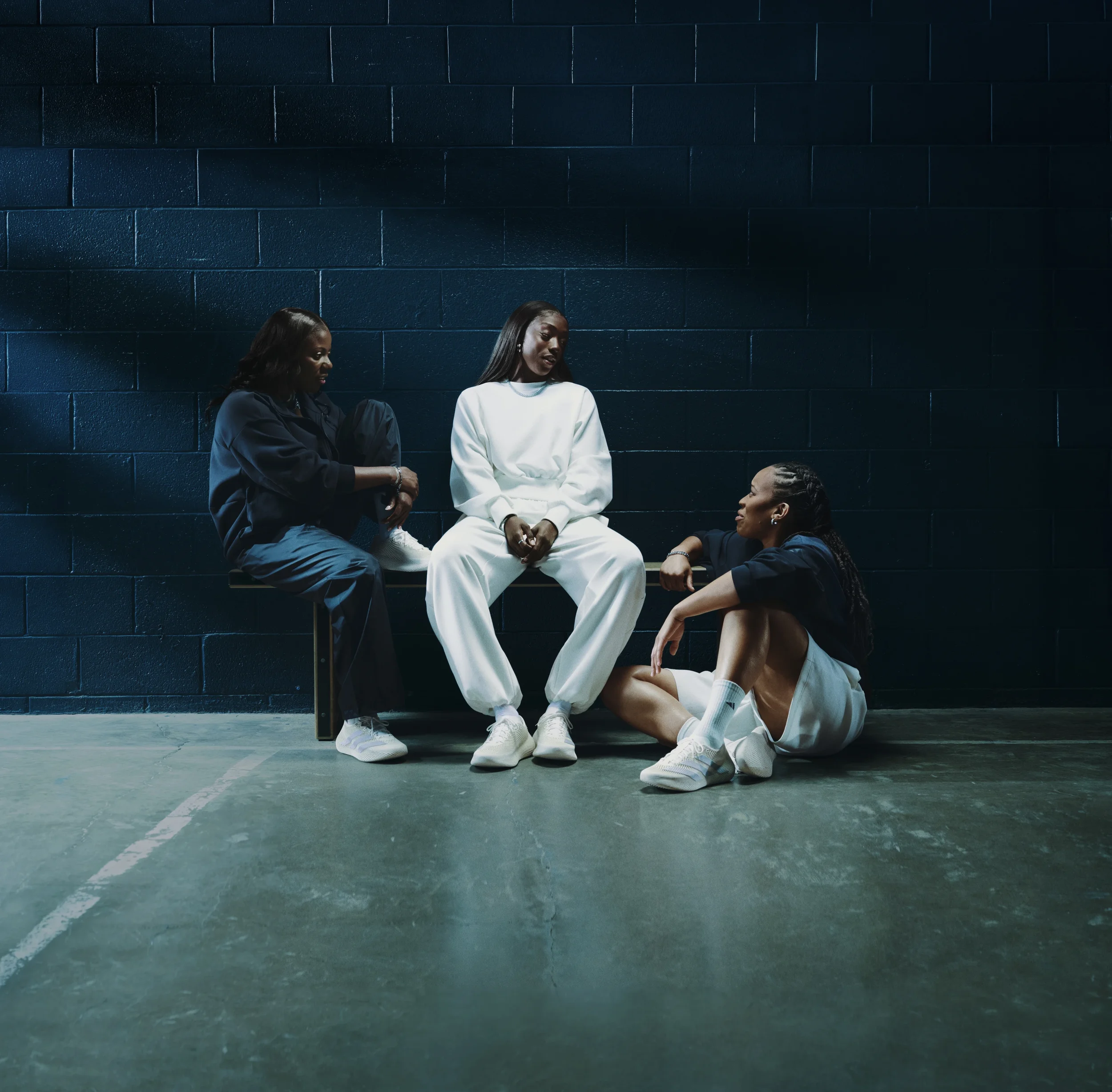 Four women with dark hair and dark skin sitting against a dark blue brick wall in a parking lot, engaging in conversation.