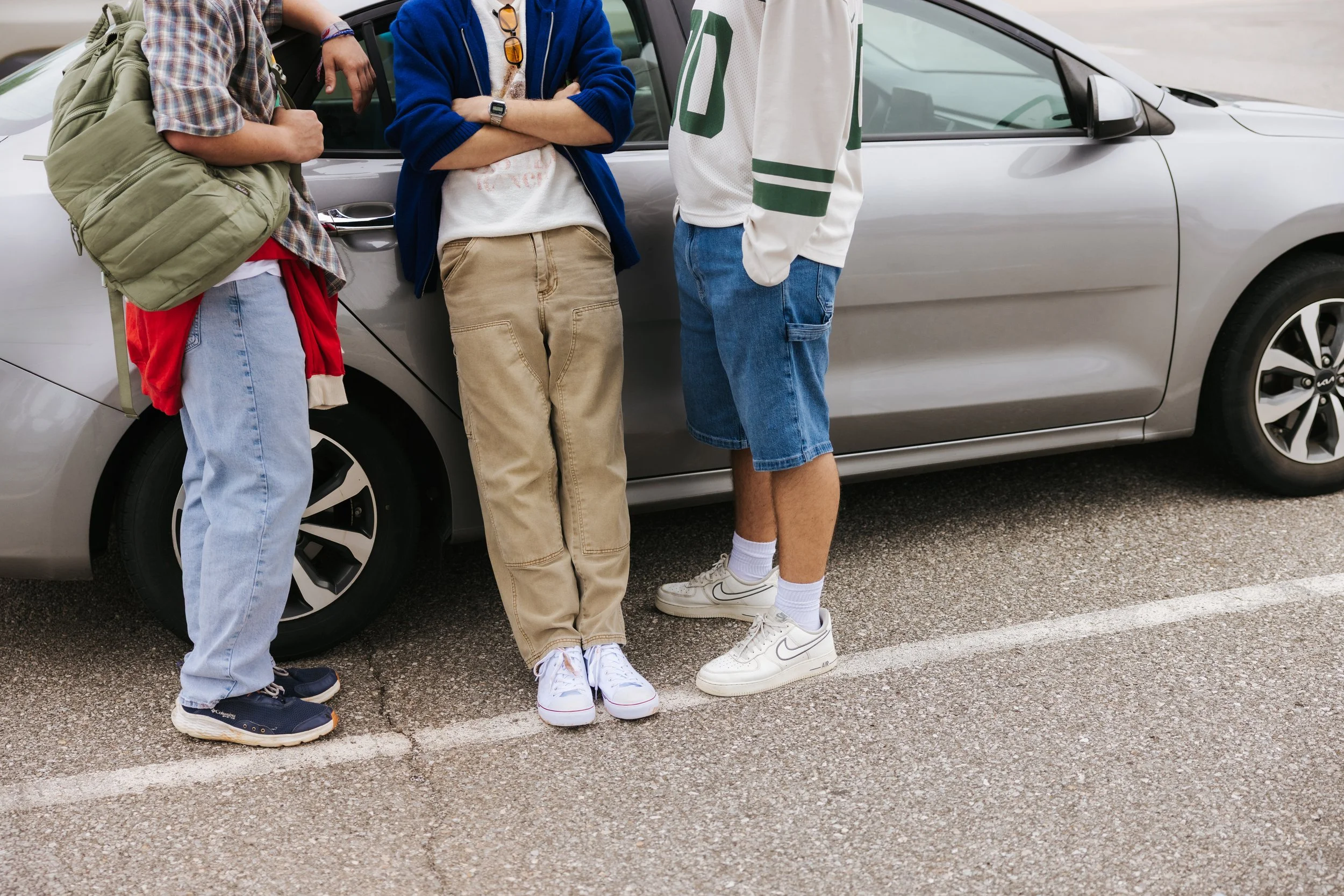 Three young people standing outside near a silver car, with partial views of their torsos and legs visible. They are dressed casually, wearing sneakers, jeans, and jackets, with backpacks and accessories.