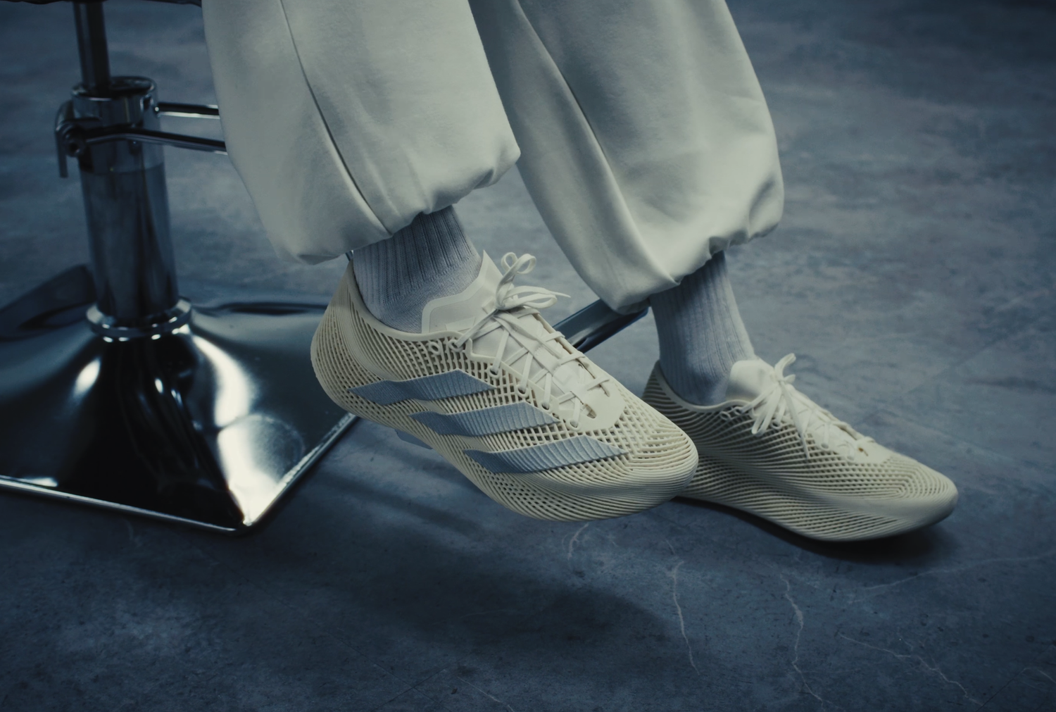 Close-up of a person wearing off-white sneakers with blue stripes, white socks, and light-colored pants, sitting on a metal stool with a chrome base on a gray concrete floor.