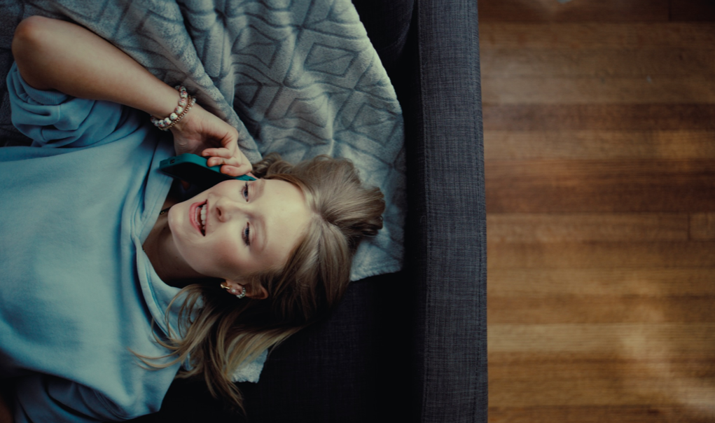 A woman lying on her back on a couch, holding a phone to her ear, smiling and looking up, with light-colored hair, wearing earrings and a light blue top.
