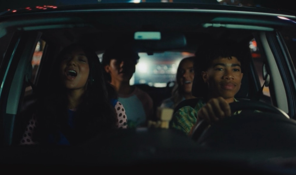 Four teenagers sitting in a car at night, with the driver smiling and the others laughing and looking in different directions.