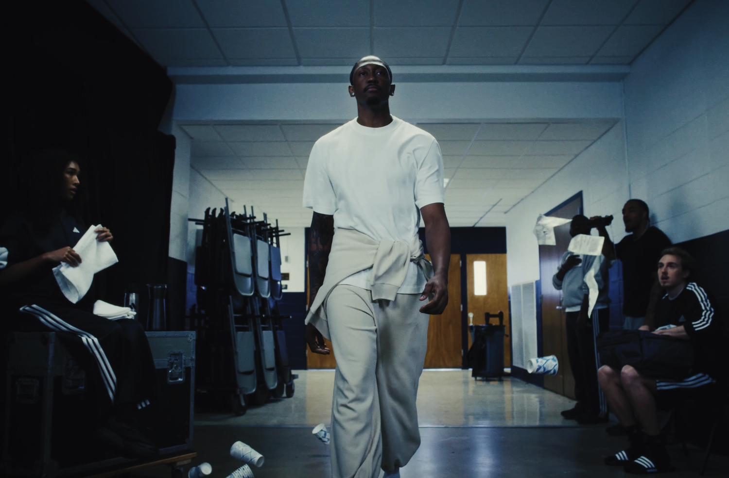 A man in a white t-shirt and beige pants walking in a locker room or backstage area, with several people seated or standing around him, some holding towels and bags, and a camera crew recording in the background.