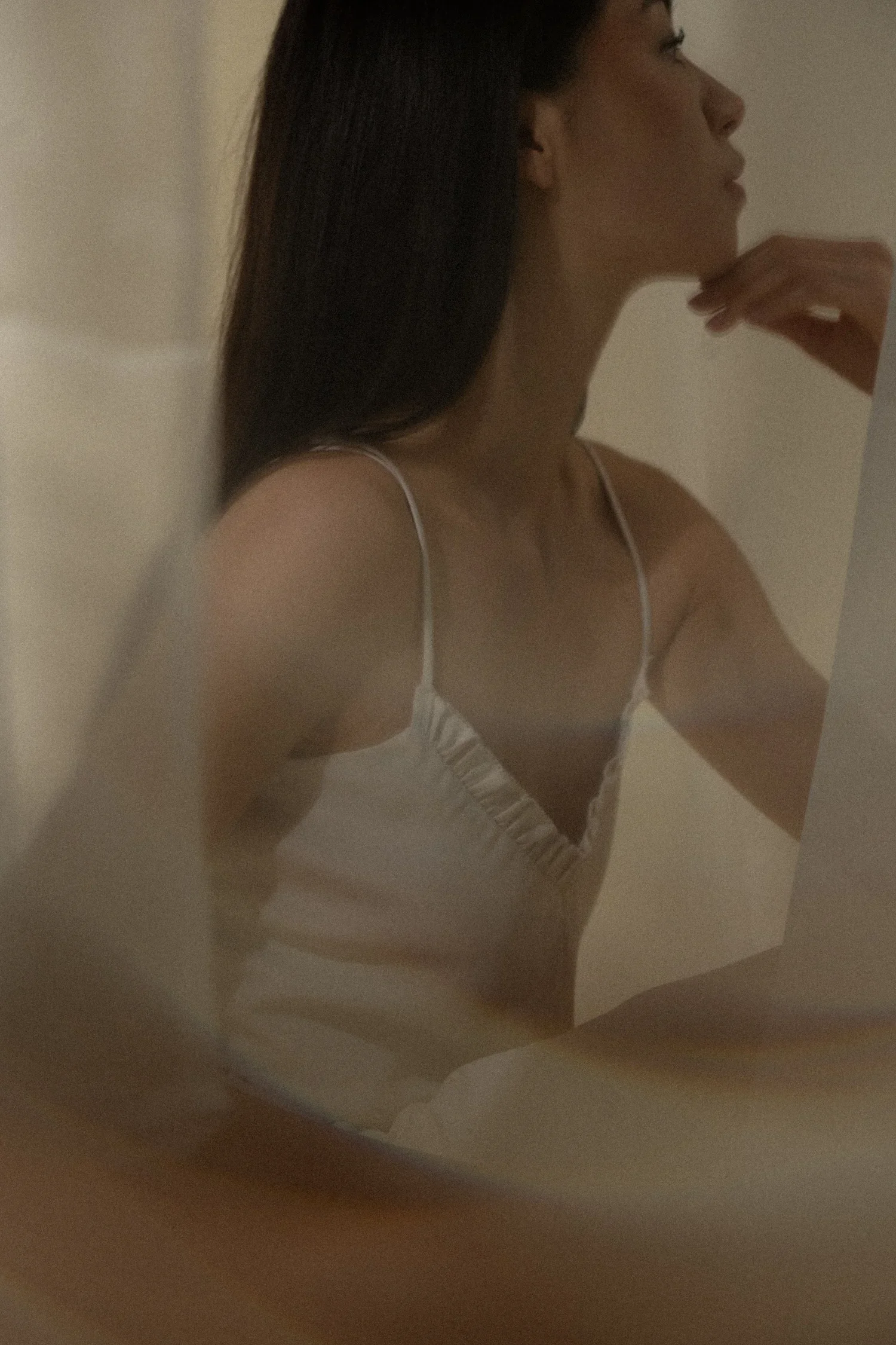A woman with long dark hair wearing a white spaghetti strap top or dress, standing in front of a mirror, with her hand near her chin, taking a selfie or a photo.