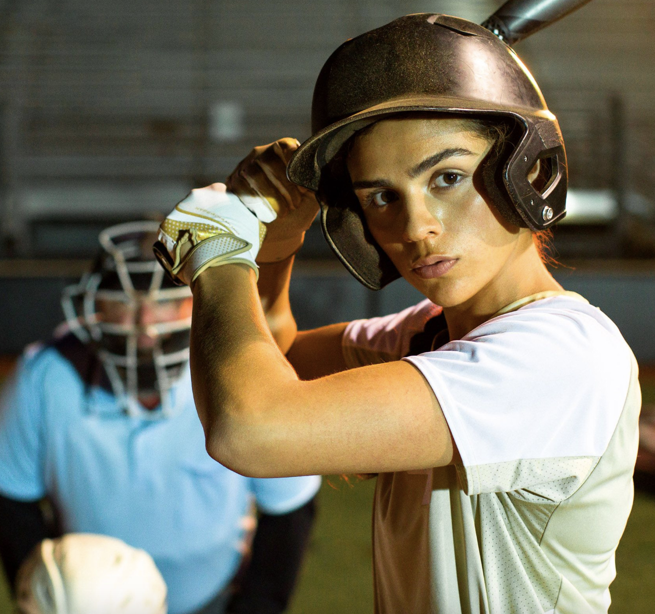 A female football player adjusting her helmet on a football field at night, with a male goalkeeper in the background.