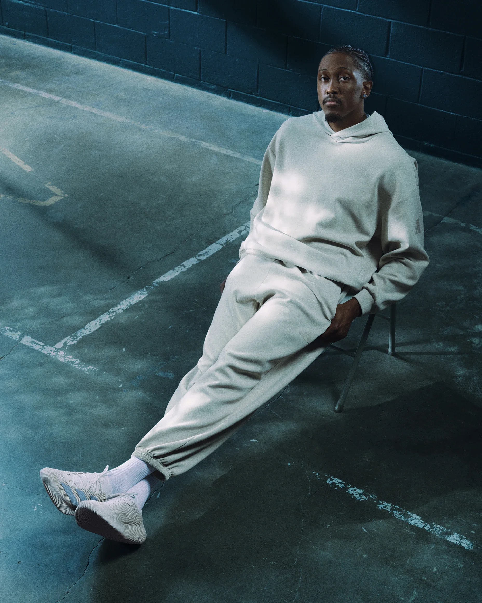 A person sitting on a chair in an indoor parking lot with a dark wall behind them, wearing a beige hoodie, matching beige pants, and white sneakers, looking at the camera.