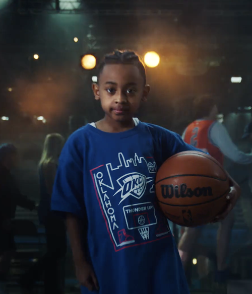 A young girl wearing a blue Oklahoma City Thunder basketball shirt holding a Wilson basketball in a dimly lit indoor basketball court.