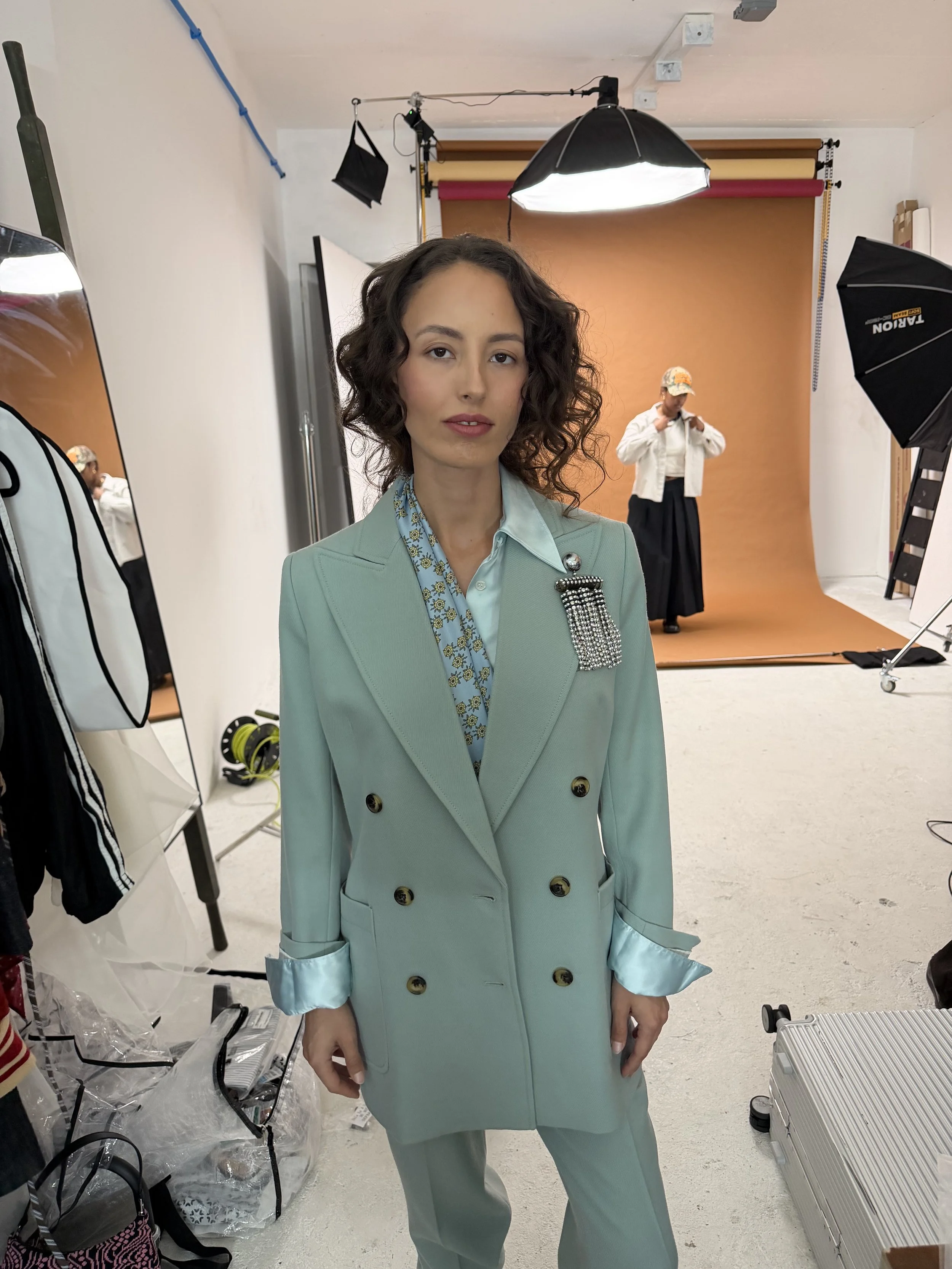 A woman in a light green suit with an ornate brooch stands in a photography studio, with a caramel-colored backdrop behind her. She has shoulder-length curly hair and a subtle smile. Behind her, a person is adjusting their clothing. Equipment like so