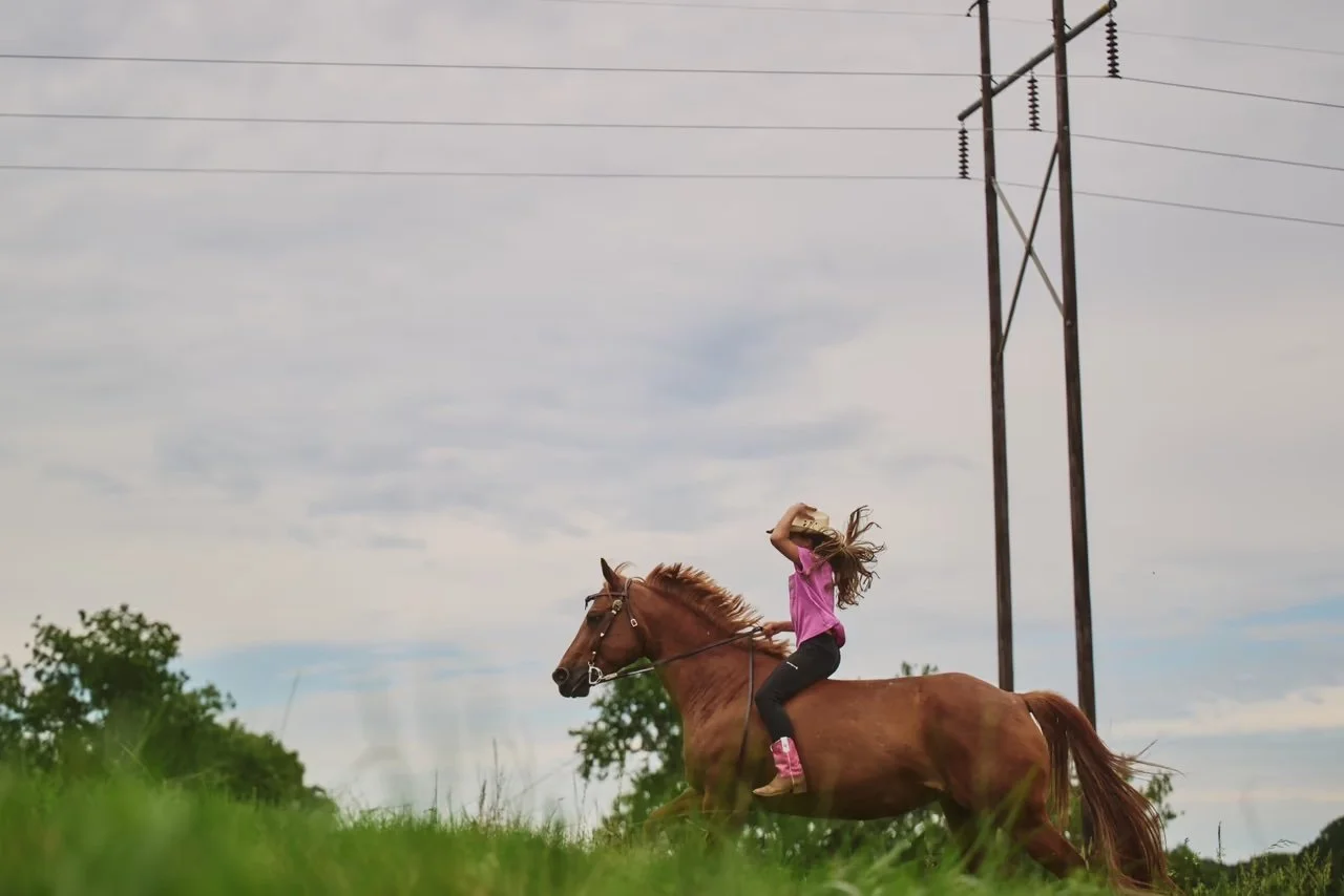 A girl riding a brown horse on a grassy field, holding a hat to her head as the horse runs, with a cloudy sky and power lines in the background.