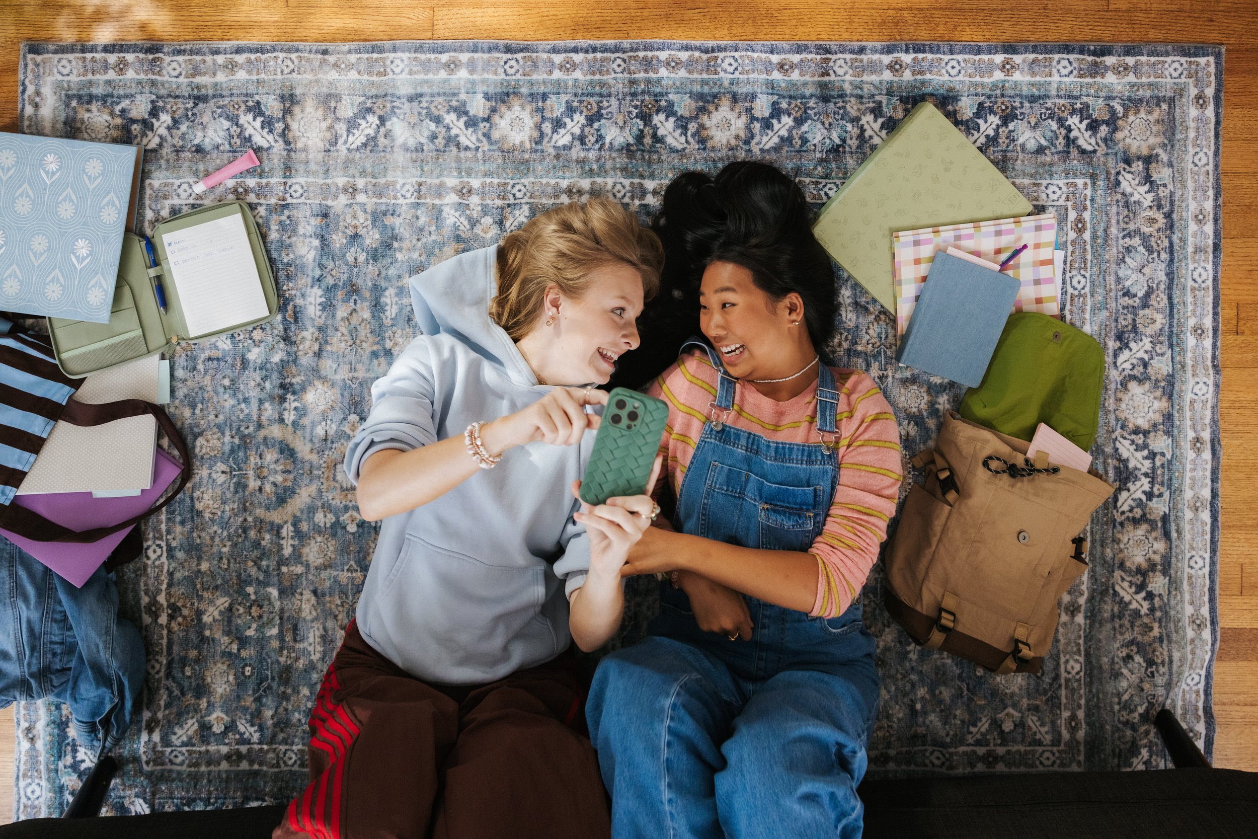 Two women lying on a patterned rug, smiling and talking while one shows her phone to the other. Surrounding them are backpacks, notebooks, and writing supplies.