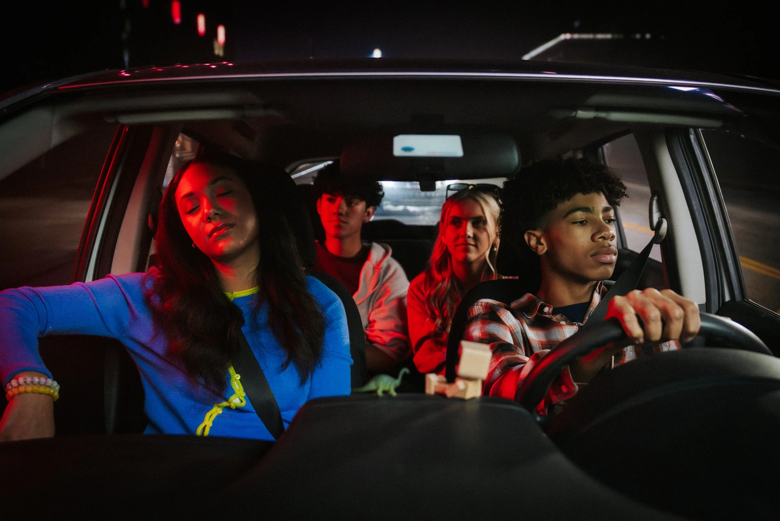 Four people are inside a car at night. The person in the driver's seat is focused on driving, while the woman in the front passenger seat has her eyes closed and appears relaxed. Two individuals are seated in the back, one with dark curly hair and th