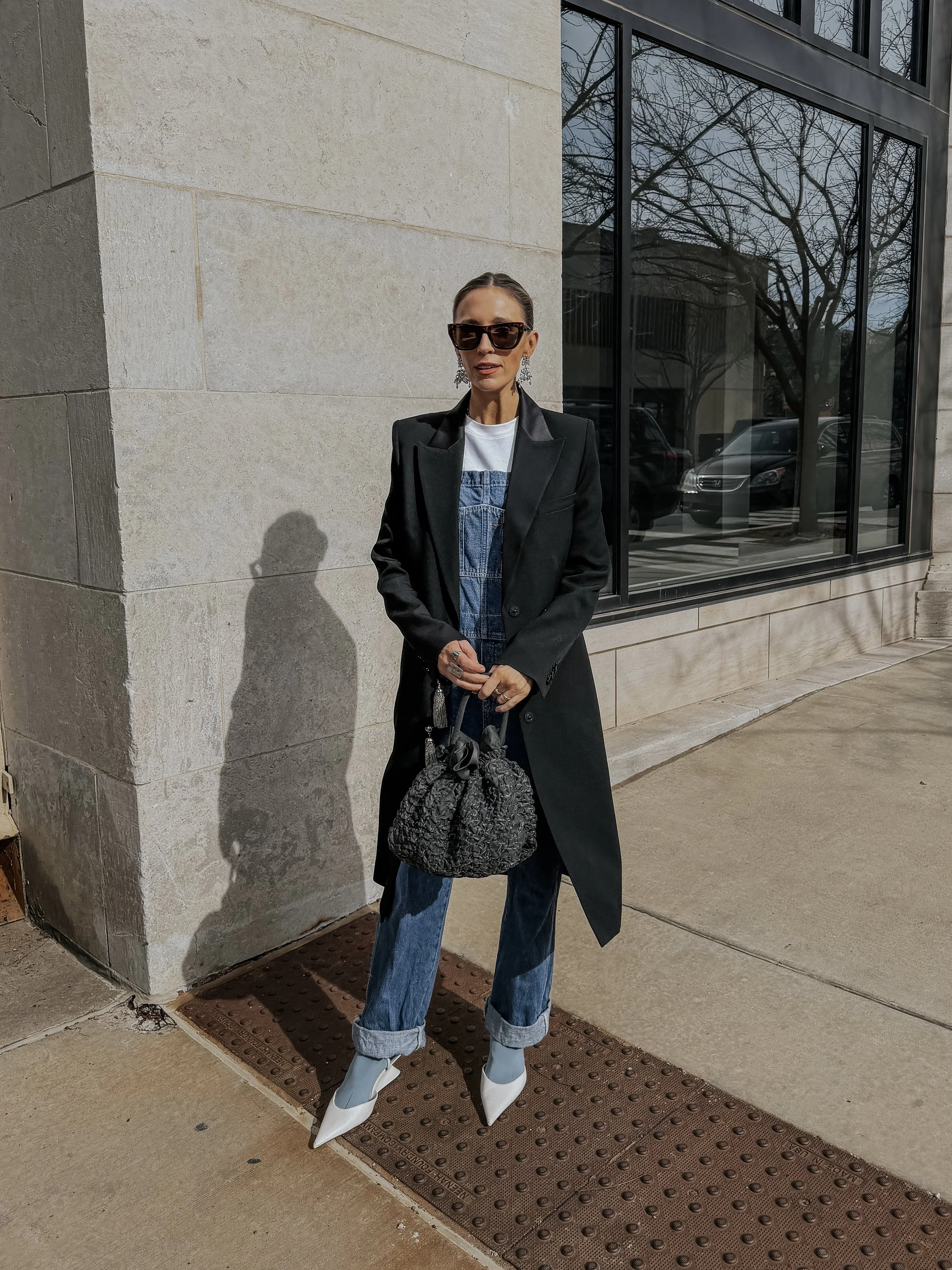 A woman with sunglasses and earrings standing outside near a building wall with large windows, wearing a black coat, denim overalls, and white shoes, holding a patterned bag.