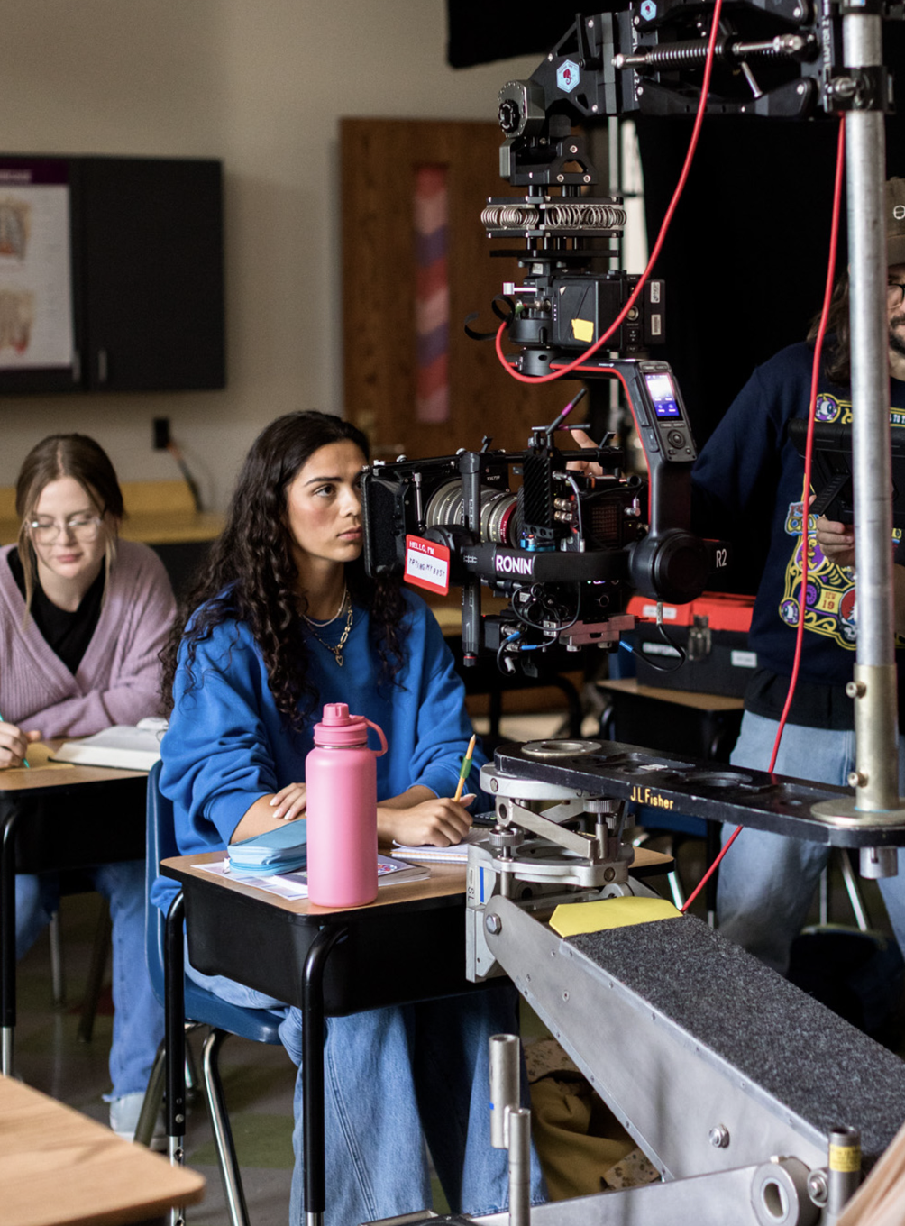 A film actress with curly dark hair and a blue sweatshirt sitting at a classroom desk, surrounded by filming equipment and a pink water bottle, while two students in the background take notes.