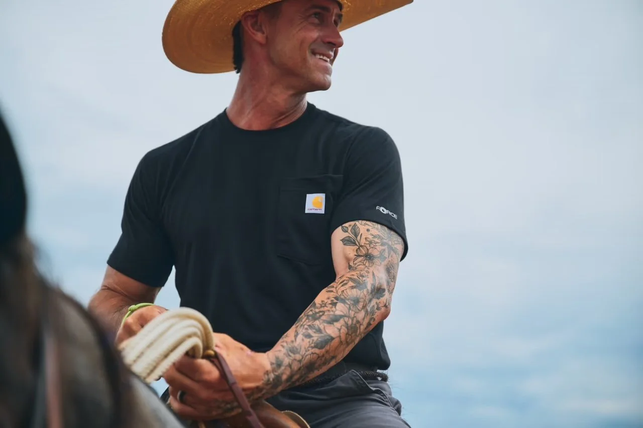 Man wearing a large straw hat and black t-shirt with a logo, smiling, holding a rope, with tattoos on his arm, outdoors with a cloudy sky background.