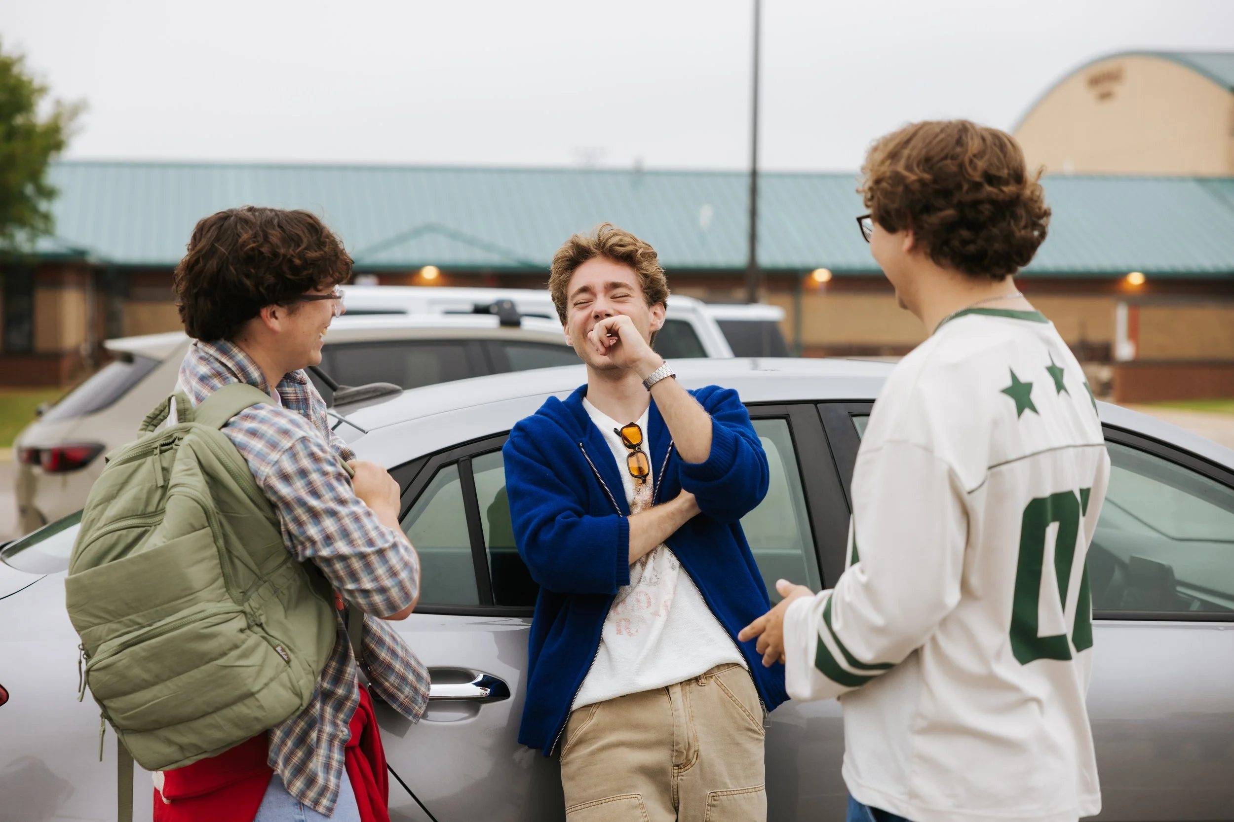 Three young men laughing and talking near a silver car in a parking lot, with a building and a cloudy sky in the background.