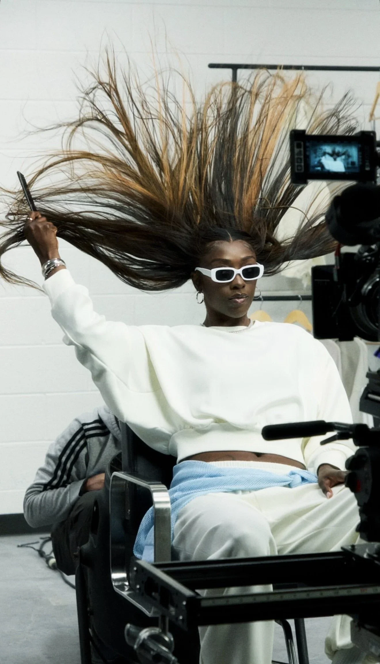 A woman with long brown hair flying in the air, wearing white sunglasses and a white sweatshirt, seated in a chair, with a camera recording her. She is in a room with a plain white wall.