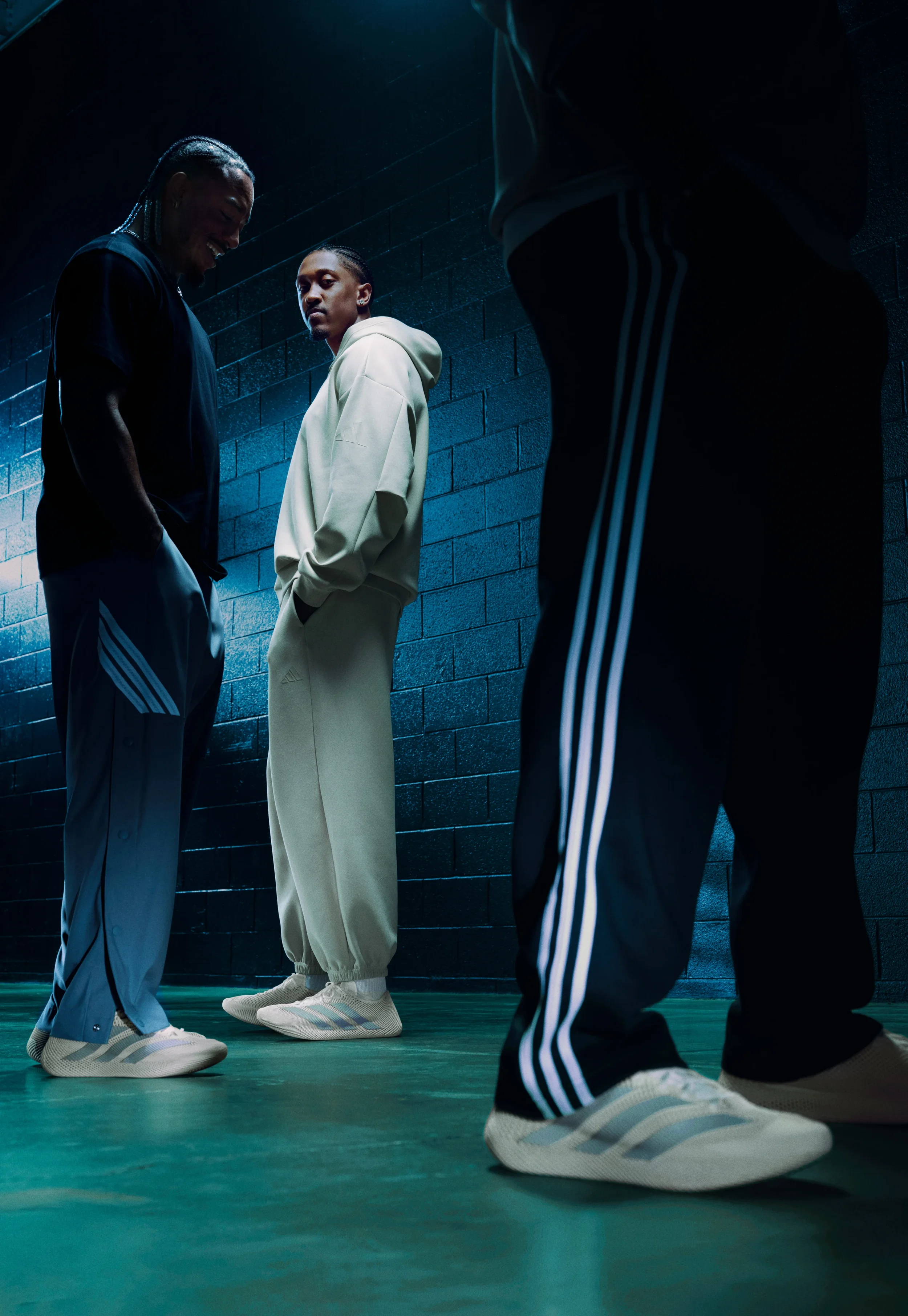 Three people in athletic clothing standing in a dimly lit indoor space with a dark brick wall, wearing sneakers, with only their lower bodies and one person's face visible.