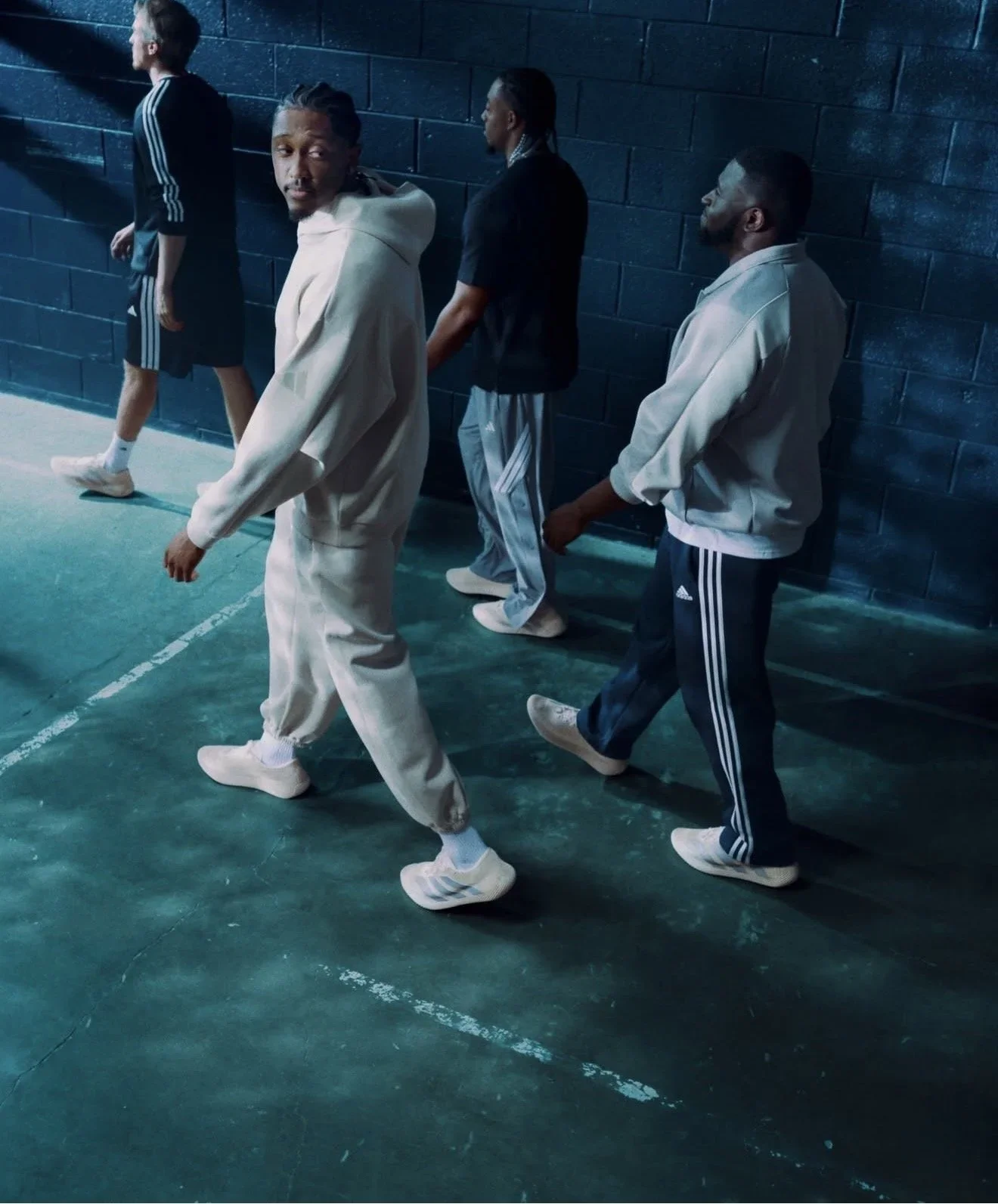 Four men standing in a row against a blue brick wall, dressed in athletic clothing and sneakers, some with hands in pockets, in a gym or sports facility with a green floor.