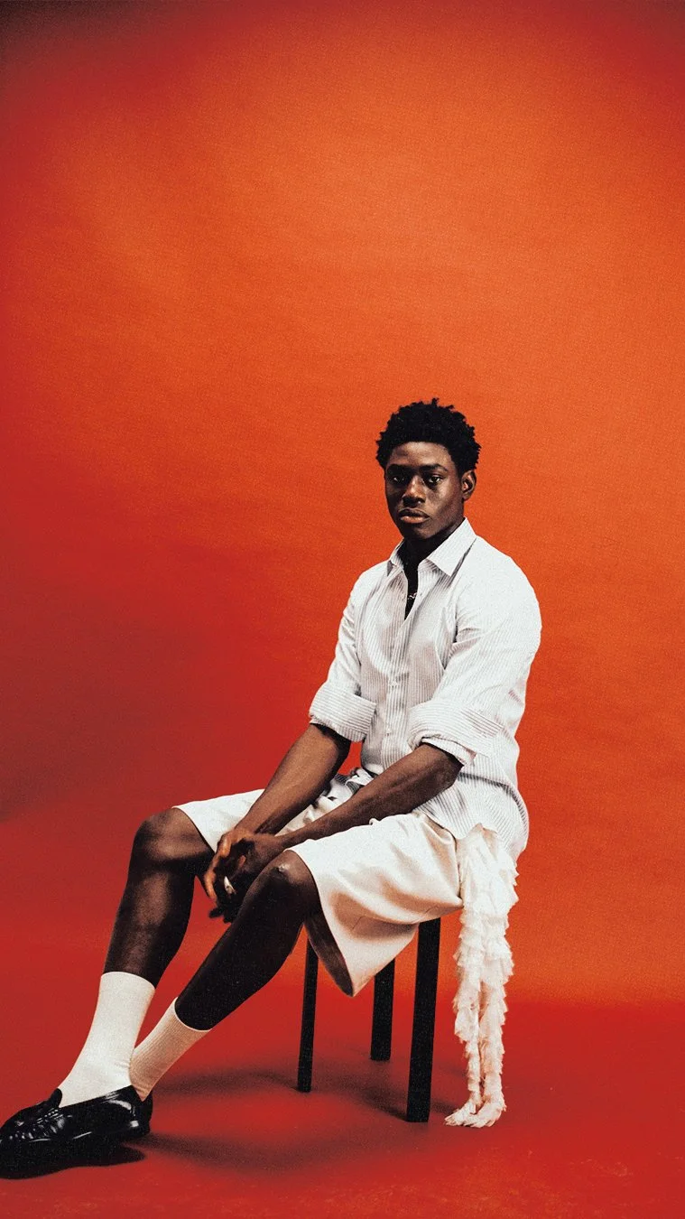 A young man with dark curly hair, wearing a white striped shirt with rolled-up sleeves, beige shorts, white socks, and black shoes, sitting on a black stool against a red-orange background.