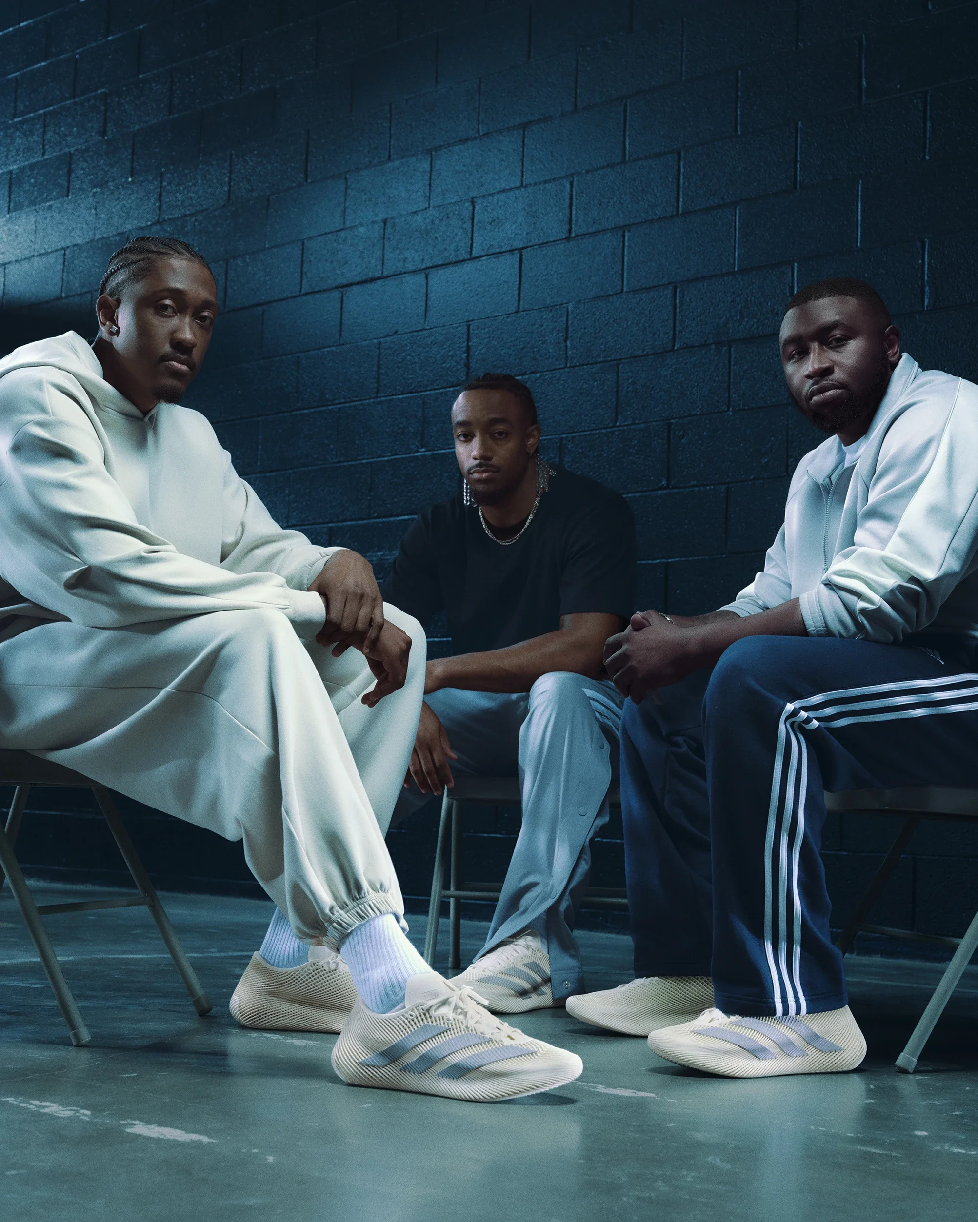 Three men sitting on chairs in front of a dark blue brick wall, wearing casual athletic clothing and matching white sneakers, with serious expressions.