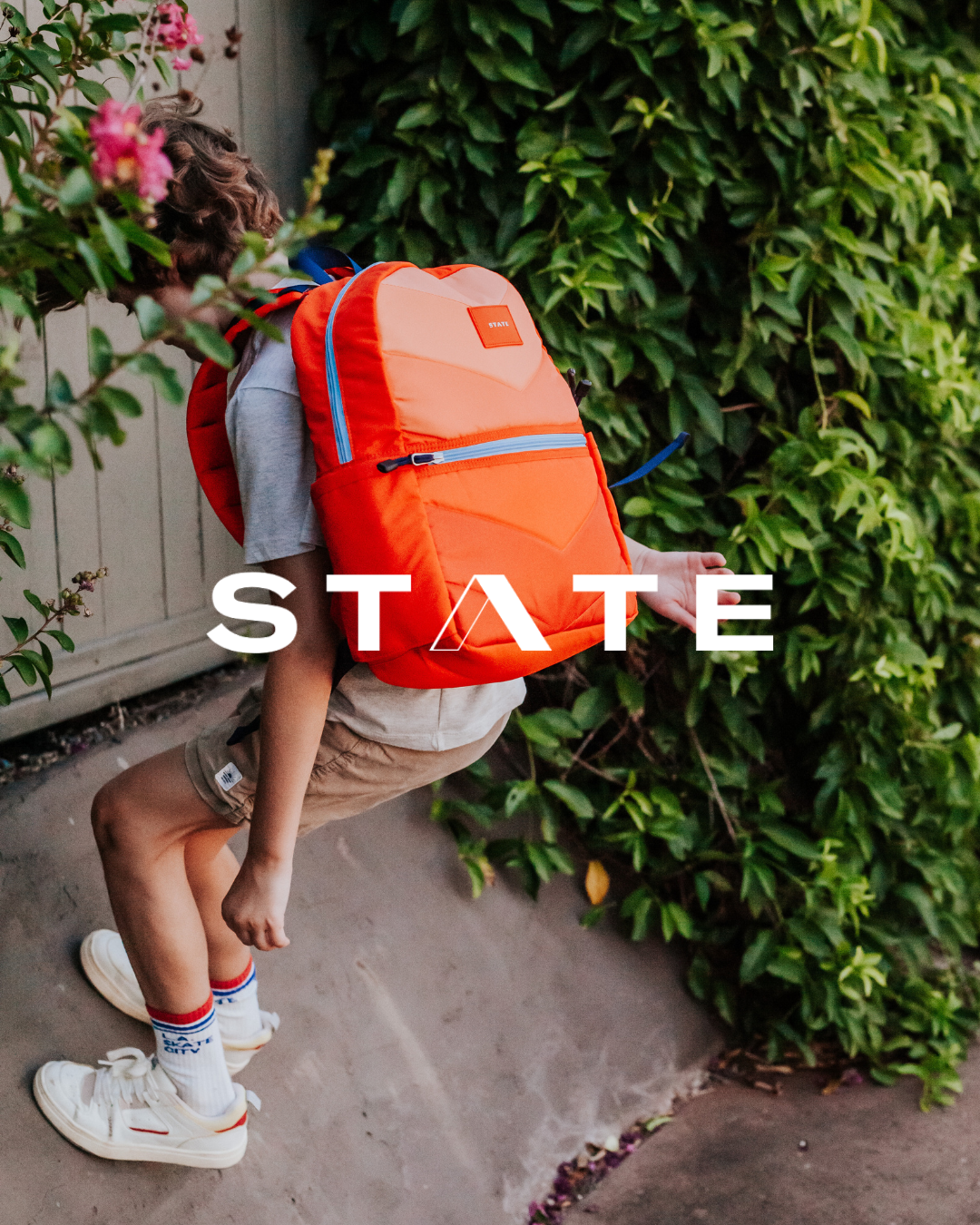 Child with curly hair wearing a gray t-shirt, beige shorts, and white sneakers, carrying an orange backpack while leaning against a fence and greenery.