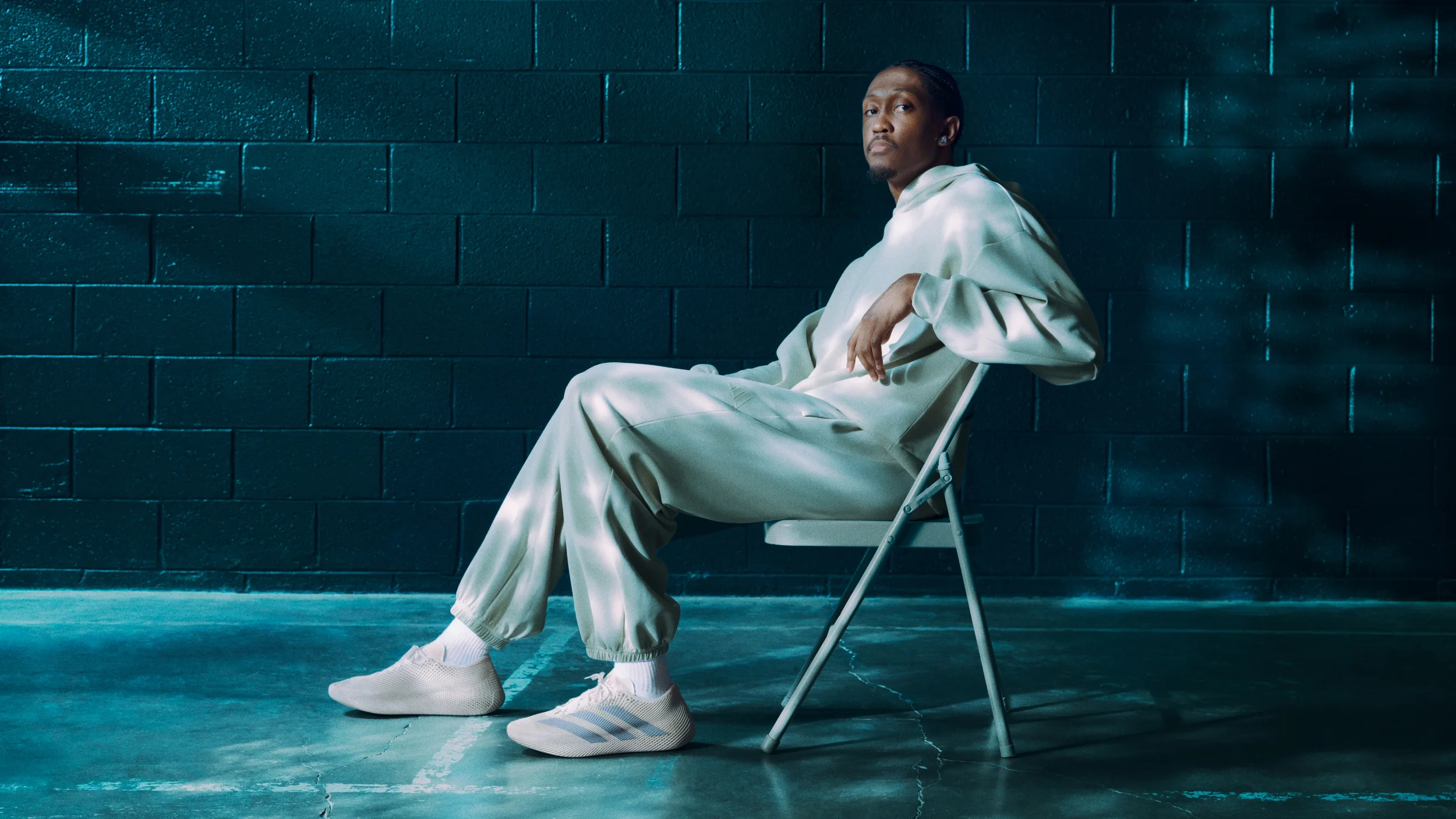 A young man wearing a shiny beige tracksuit and white sneakers sitting on a folding chair against a dark teal cinder block wall.
