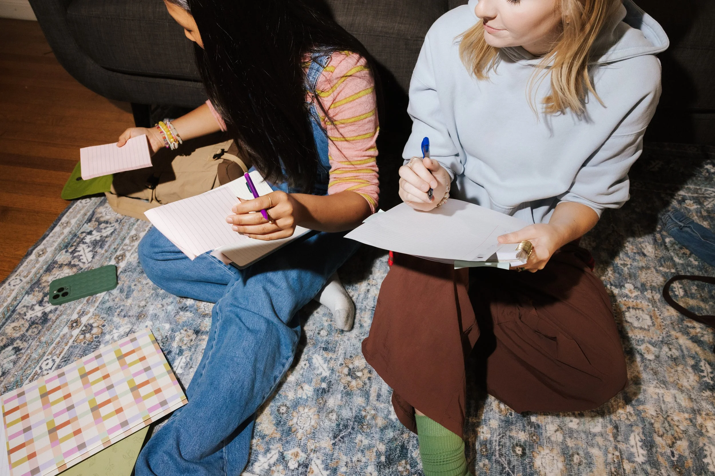 Two women sitting on a patterned rug, writing on notepads. One woman has long dark hair, wearing colorful bracelets and a striped shirt. The other woman has blonde hair, wearing a white hoodie, brown pants, and green socks. There are notebooks, a green phone case, and a colorful checkered notebook on the floor.
