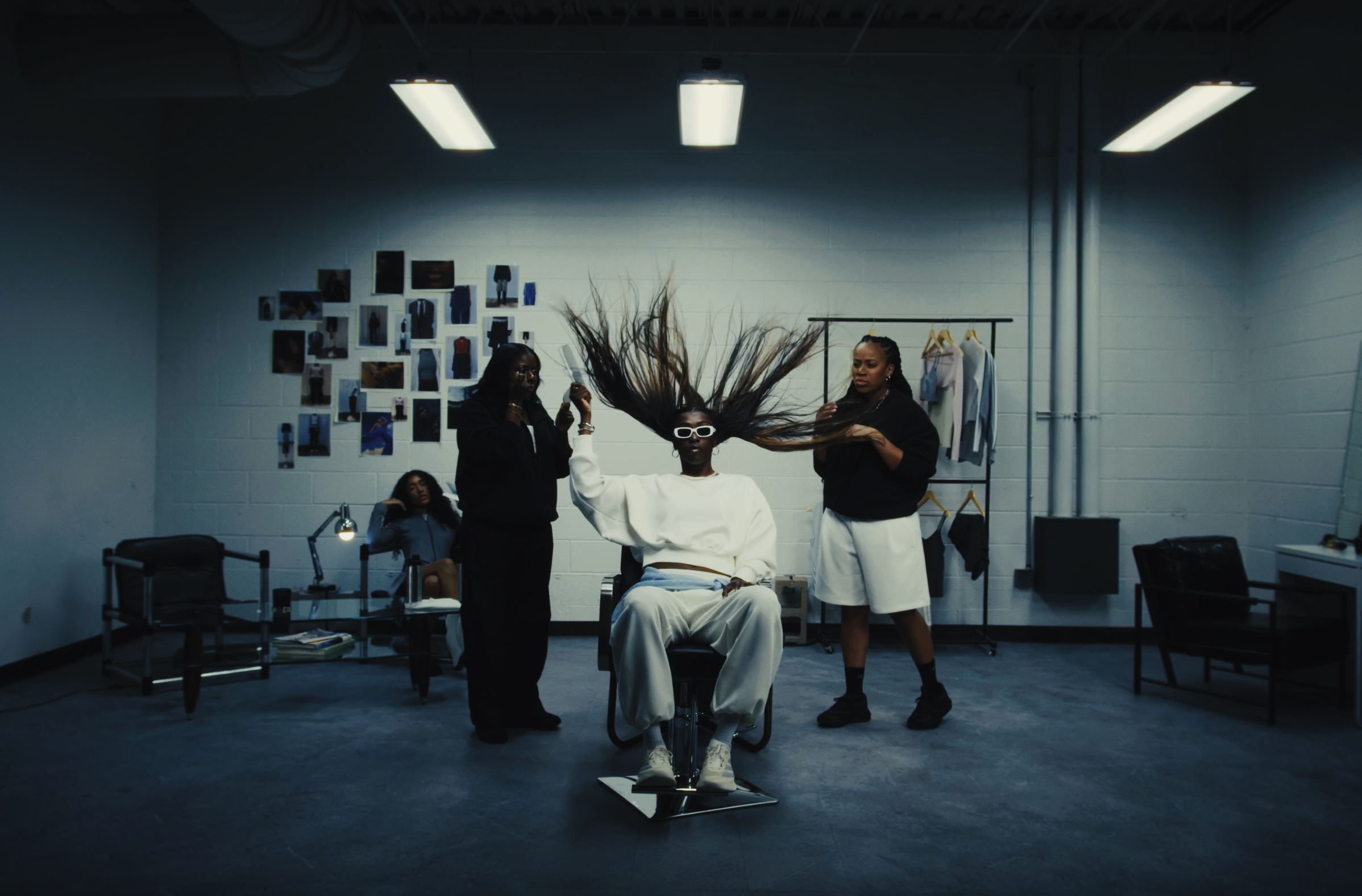 A woman with long hair wearing sunglasses sits in a chair in a minimalist room with three women around her. Two women are adjusting her hair, and a woman in the background is sitting at a desk. The room has white walls decorated with photos, a clothing rack, and fluorescent ceiling lights.