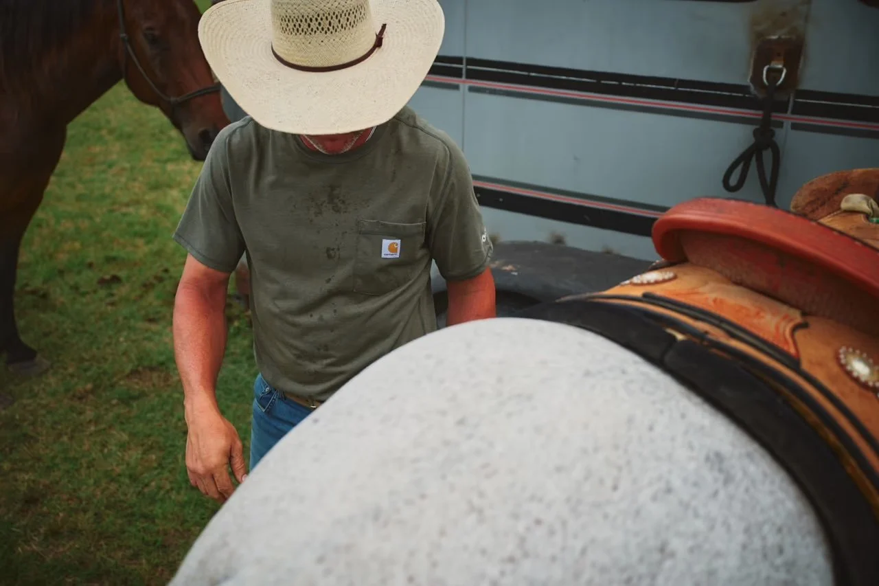 A person wearing a large straw hat, a green Carhartt T-shirt, and blue jeans, standing next to a horse with a saddle, on a grassy area.