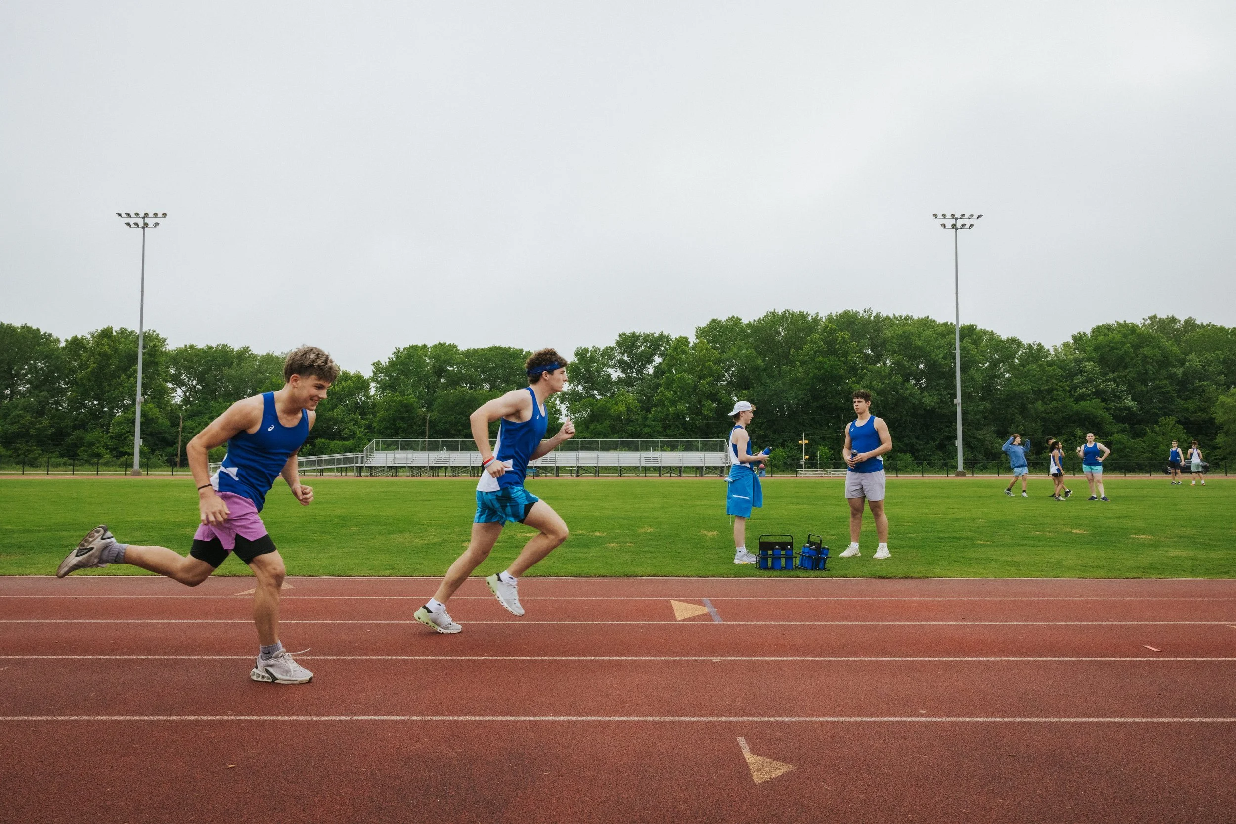 Young male athletes in blue uniforms running on a track during a race at an outdoor sports stadium, with a few officials and other athletes in the background under a cloudy sky.