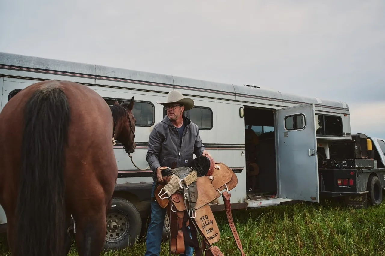 A man wearing a cowboy hat and glasses stands near a brown horse and a large white horse trailer. He holds a saddle in one hand and is dressed in a gray jacket. The setting appears to be outdoors on a grassy field.