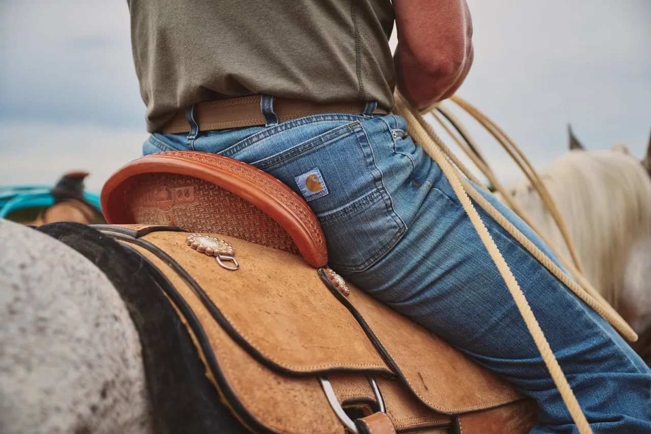 Close-up of a person riding a horse, wearing blue jeans and a brown belt, with a saddle and braided reins visible.
