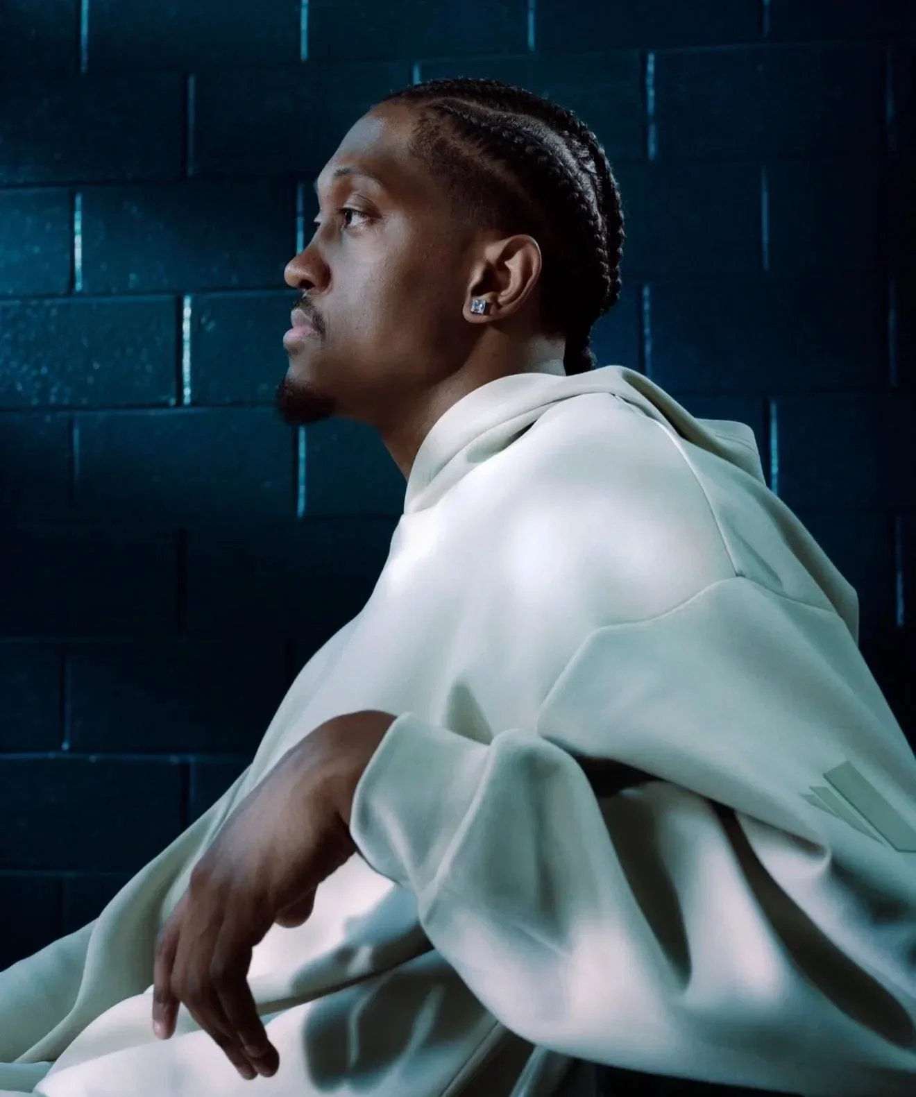 Side profile of a man with braided hair, wearing diamond earrings and a light-colored hoodie, sitting against a dark blue brick wall.