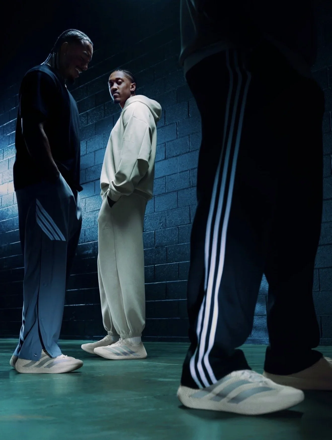 Three young men in athleticwear standing in a dimly lit studio with a concrete wall background, dressed in casual sports clothing and sneakers, engaging in a conversation or waiting.