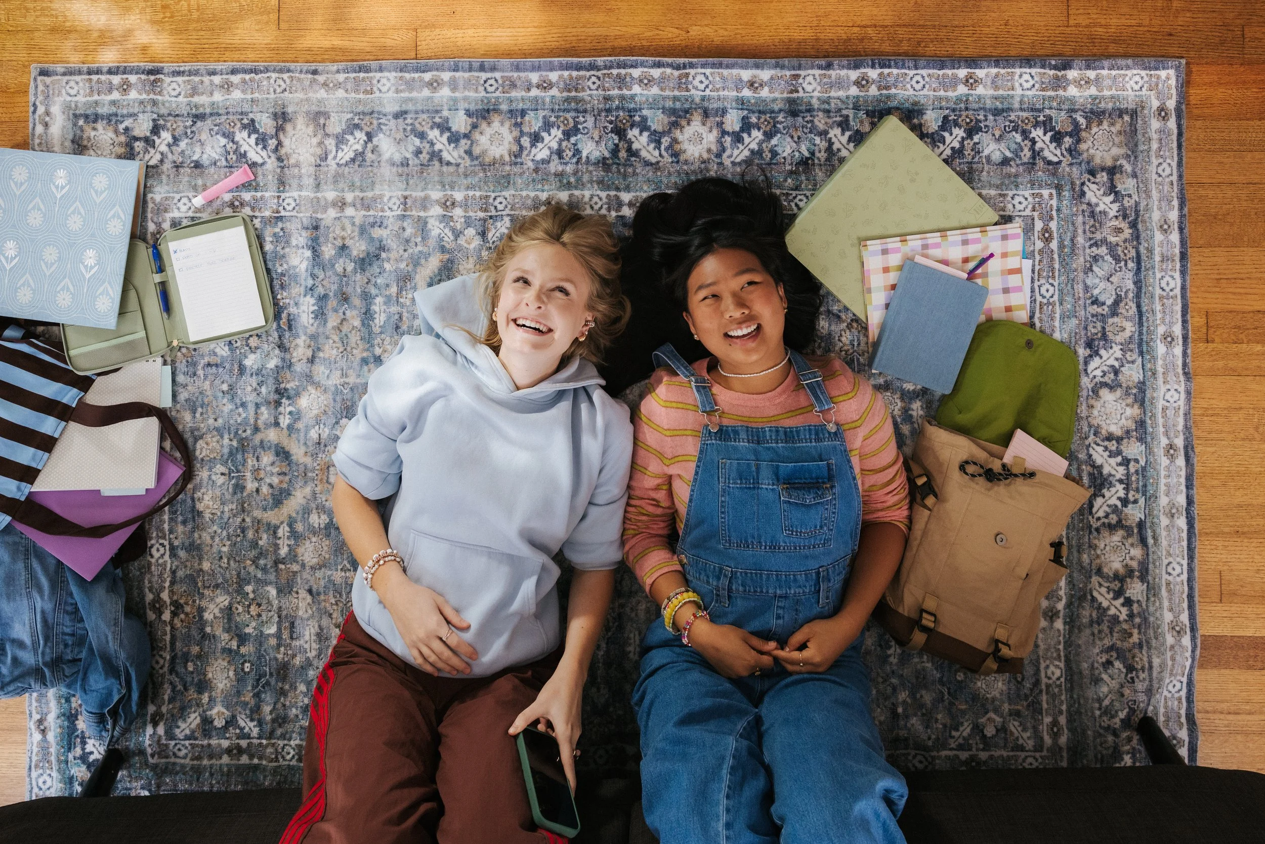 Two women lying on a blue patterned rug, smiling and looking up, surrounded by books, notebooks, backpacks, and personal items.