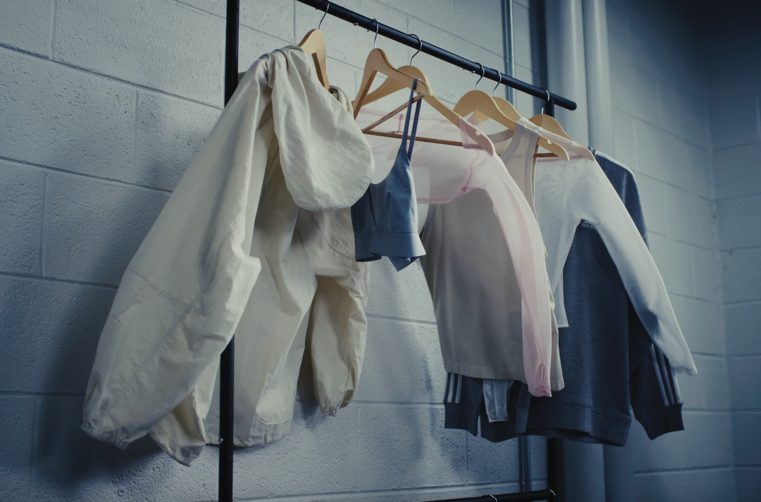 Clothes, including a jacket, a face mask, a sports bra, a t-shirt, a sweatshirt, and a hoodie, hanging on a clothing rack against a white brick wall.