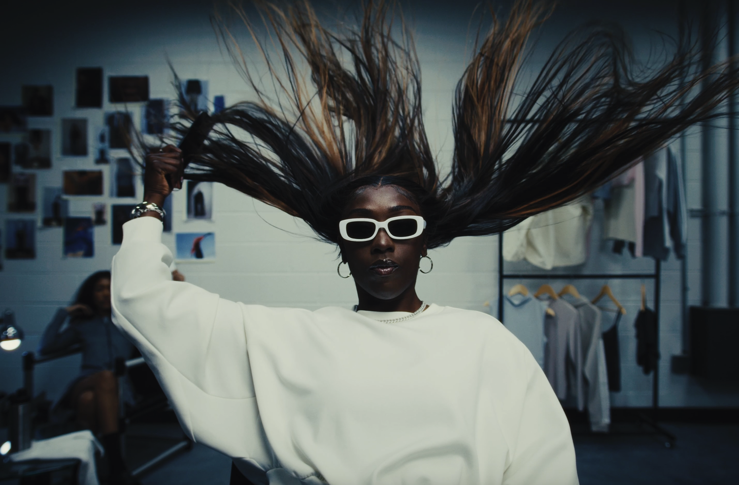 Fashionably styled woman with long flowing hair, wearing white sunglasses and hoodie, posing indoors with a gallery wall of photos behind her.