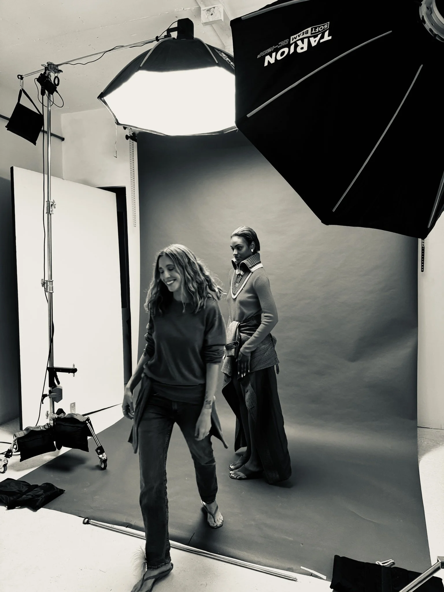 Black and white photo of two women in a photography studio. One woman with long wavy hair is smiling and walking, while the other with short hair is standing in the background, gazing. Photography equipment, including softboxes, a light stand, and a backdrop, surrounds them.
