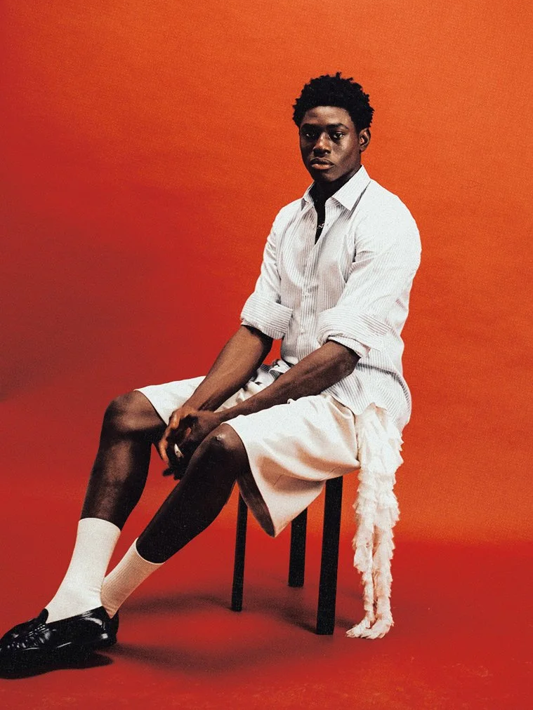 A young man with dark skin, short curly hair, and a serious expression, seated on a black stool against an orange background. He is wearing a white striped button-up shirt with rolled-up sleeves, light-colored shorts, white socks, and black shoes, with a white fluffy scarf hanging from the stool.
