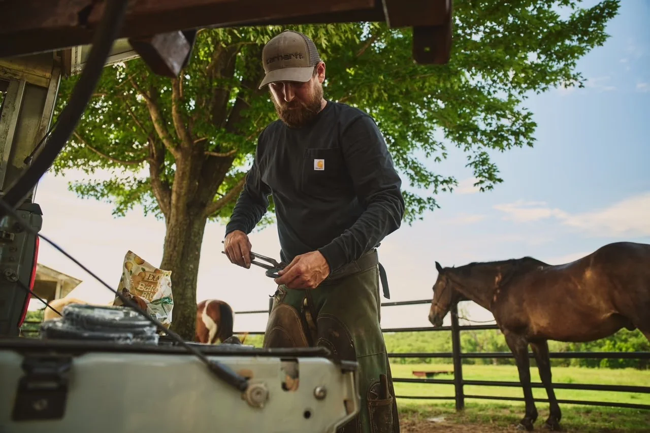 A bearded man wearing a baseball cap and dark long sleeve shirt preparing food outdoors, with a horse and trees in the background.