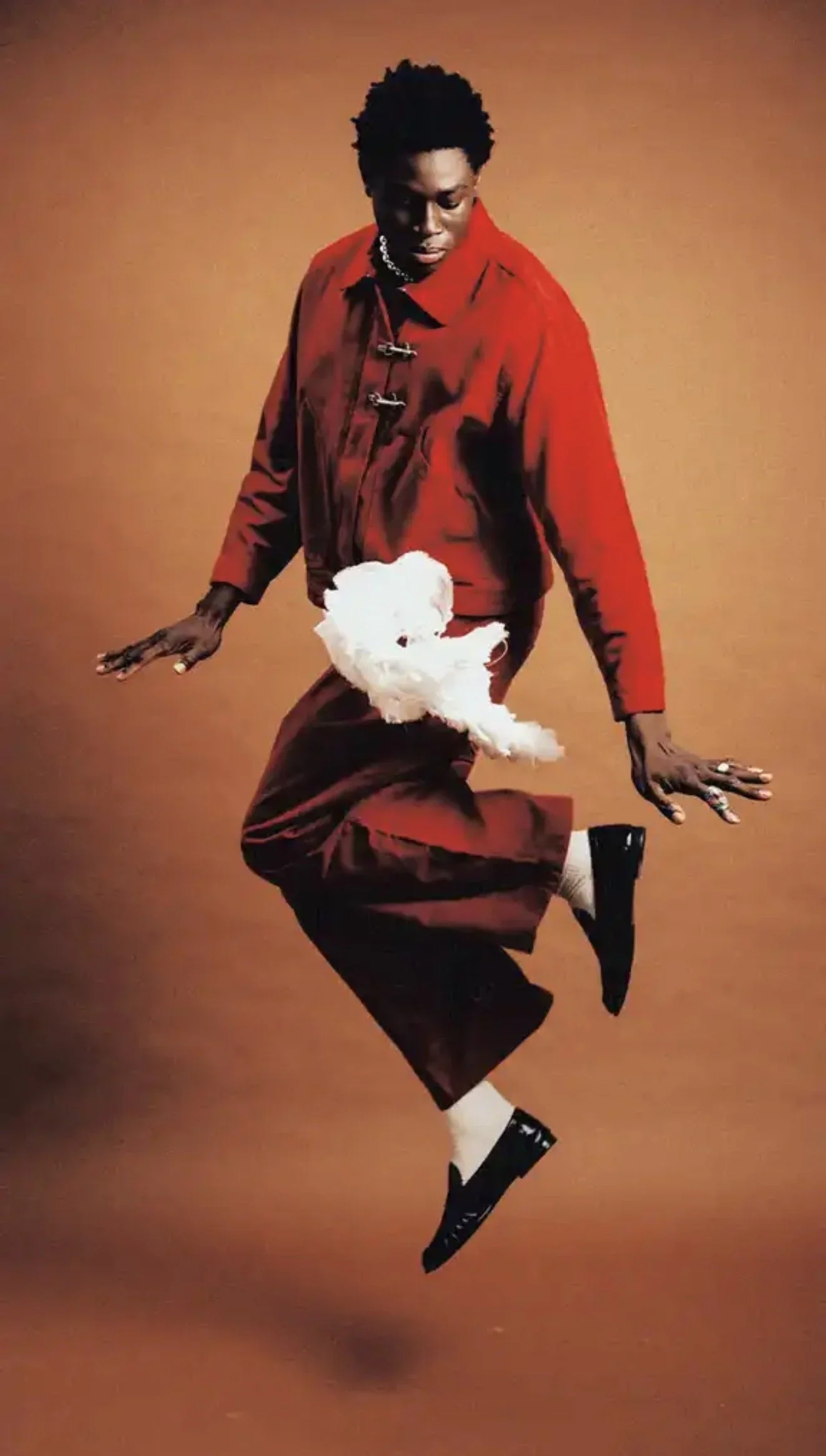 A person with dark skin and short curly hair is mid-air against a plain brown background, wearing a red jacket, maroon pants, and black shoes, with a fluffy white dog in front of them.